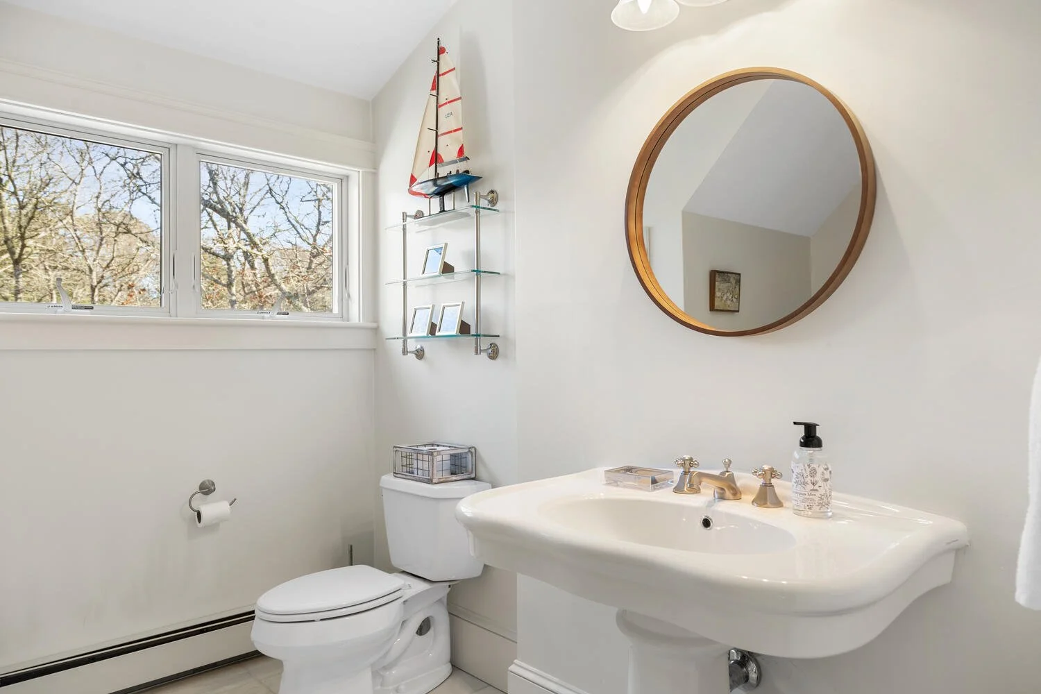 Small bathroom with a white toilet, a white sink, and a round mirror. There is a window with outside trees, a wall shelf with a model sailboat and picture frames. A soap dispenser is on the sink.