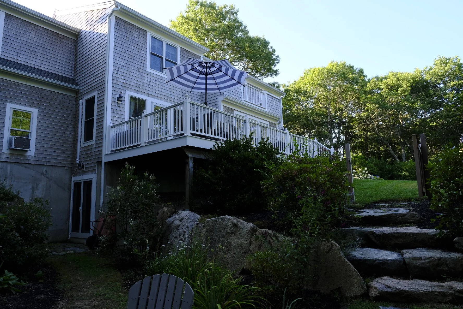 Backyard view of a house with a wooden deck featuring a striped patio umbrella, surrounded by trees, bushes, and a stone staircase leading to a grassy area.