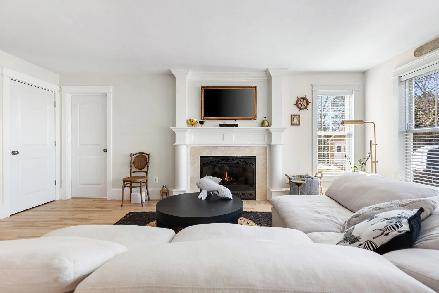 Living room with a white sofa, black round coffee table with a decorative bird sculpture, fireplace with a TV mounted above, wooden chair, windows with white blinds, plants, and modern decor.
