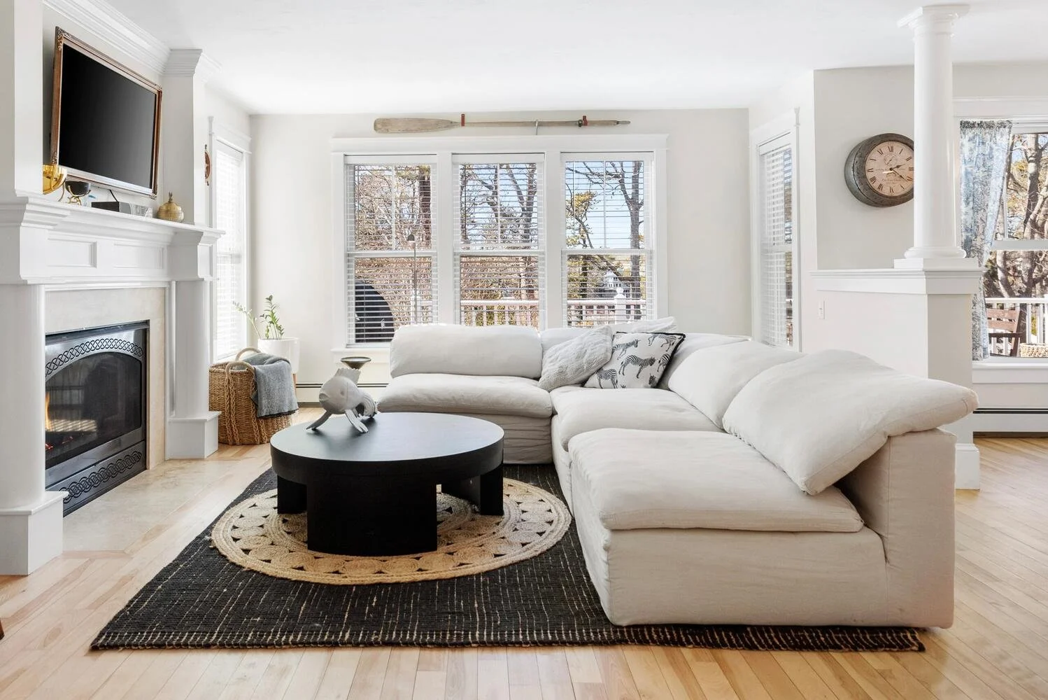Living room with white L-shaped sofa, black circular coffee table on a patterned rug, fireplace, large window with blinds, and wooden flooring.