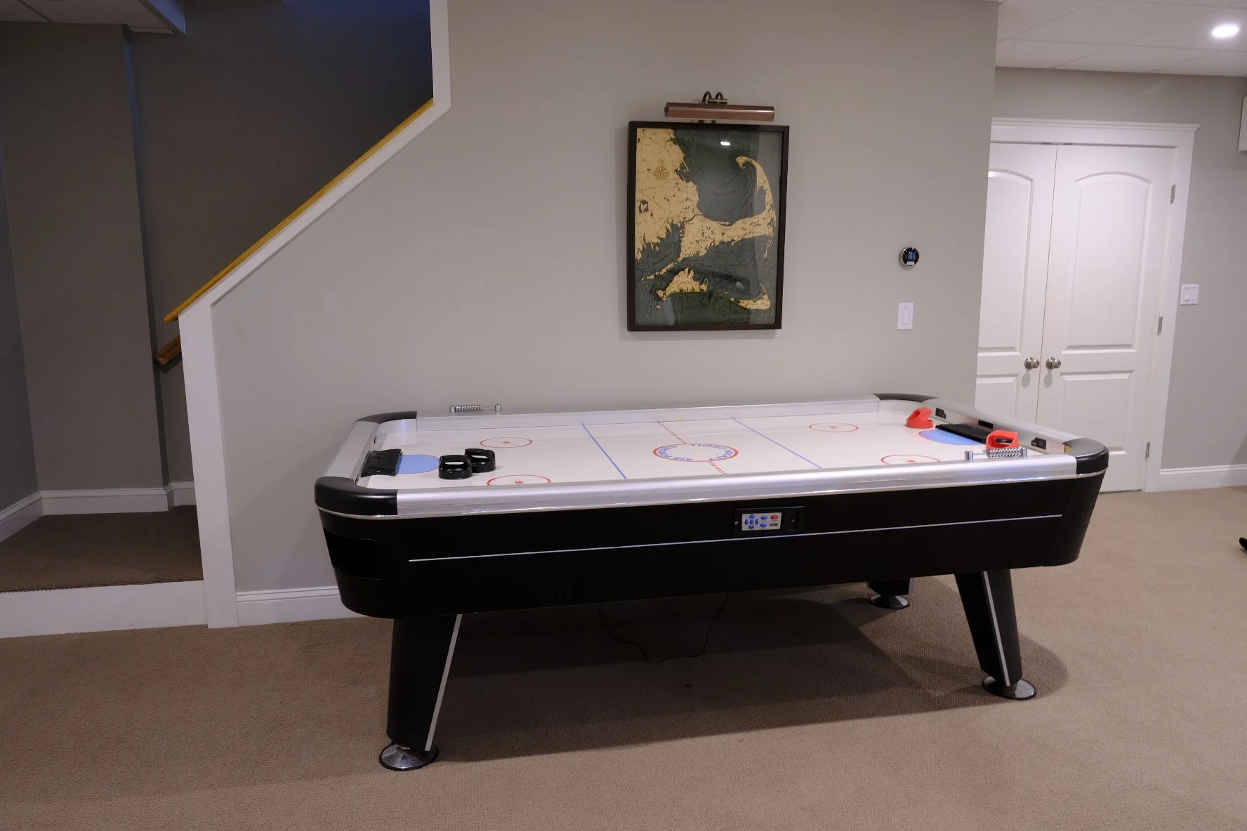 An air hockey table in a room with beige carpet, white walls, and a staircase on the left. There is framed artwork on the wall behind the table and a closed double door on the right.