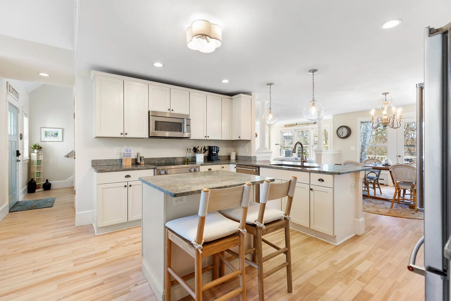 Kitchen with white cabinets, granite countertops, stainless steel appliances, a small island with two chairs, pendant lighting, and an open dining area with chandeliers and large windows.