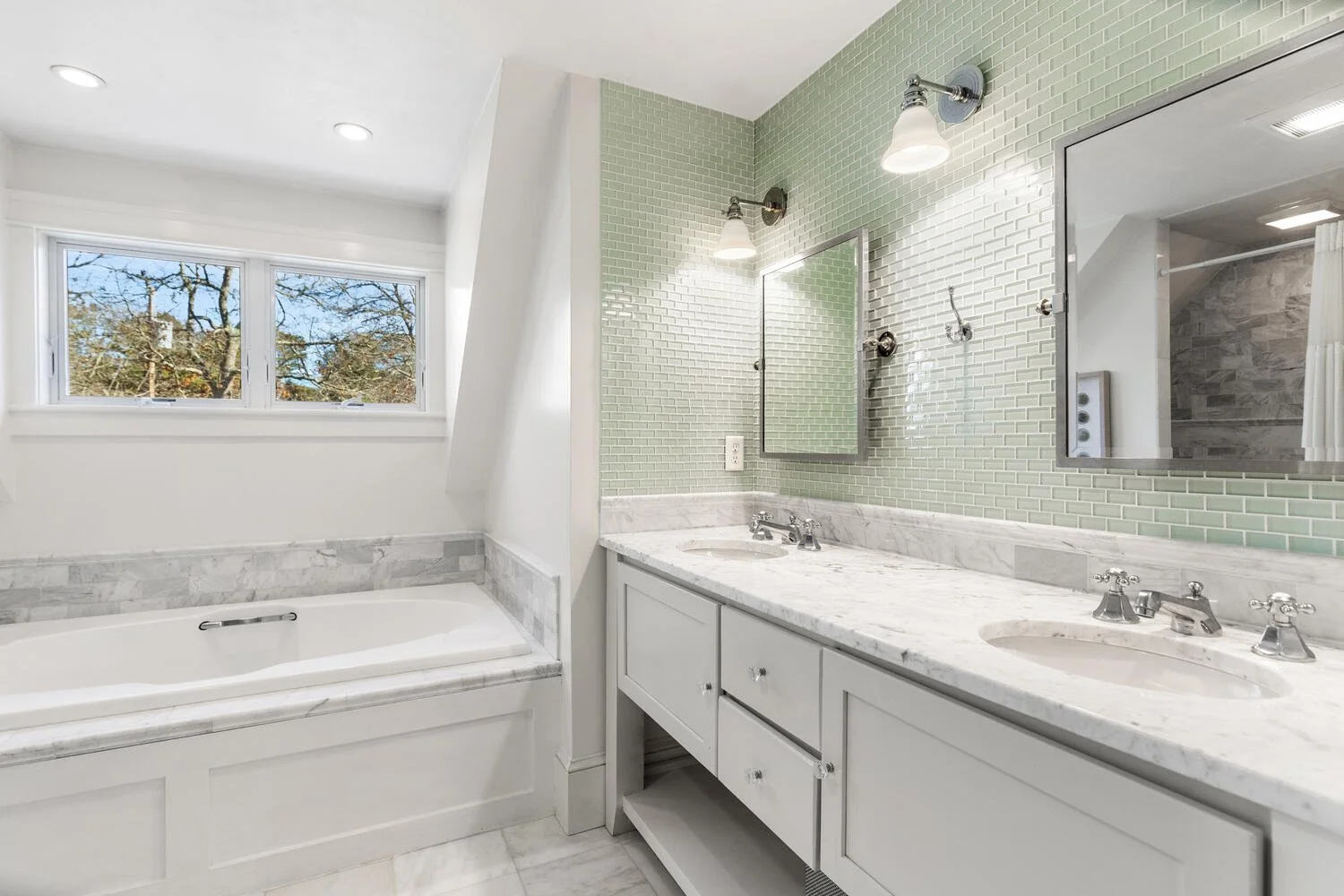 A bathroom with a double sink vanity with a marble countertop, a mirror, and green mosaic tile walls. There is a small window showing trees outside and a bathtub with marble trim.