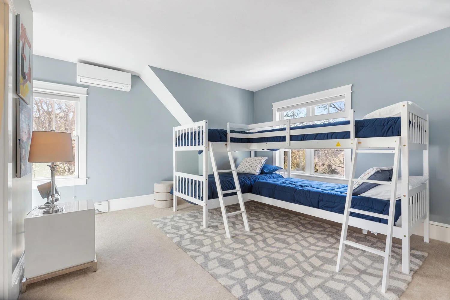 Room with light blue walls, a white bunk bed with navy bedding, a beige patterned rug, a window with tree view, a white side table with a lamp, and an air conditioning unit.