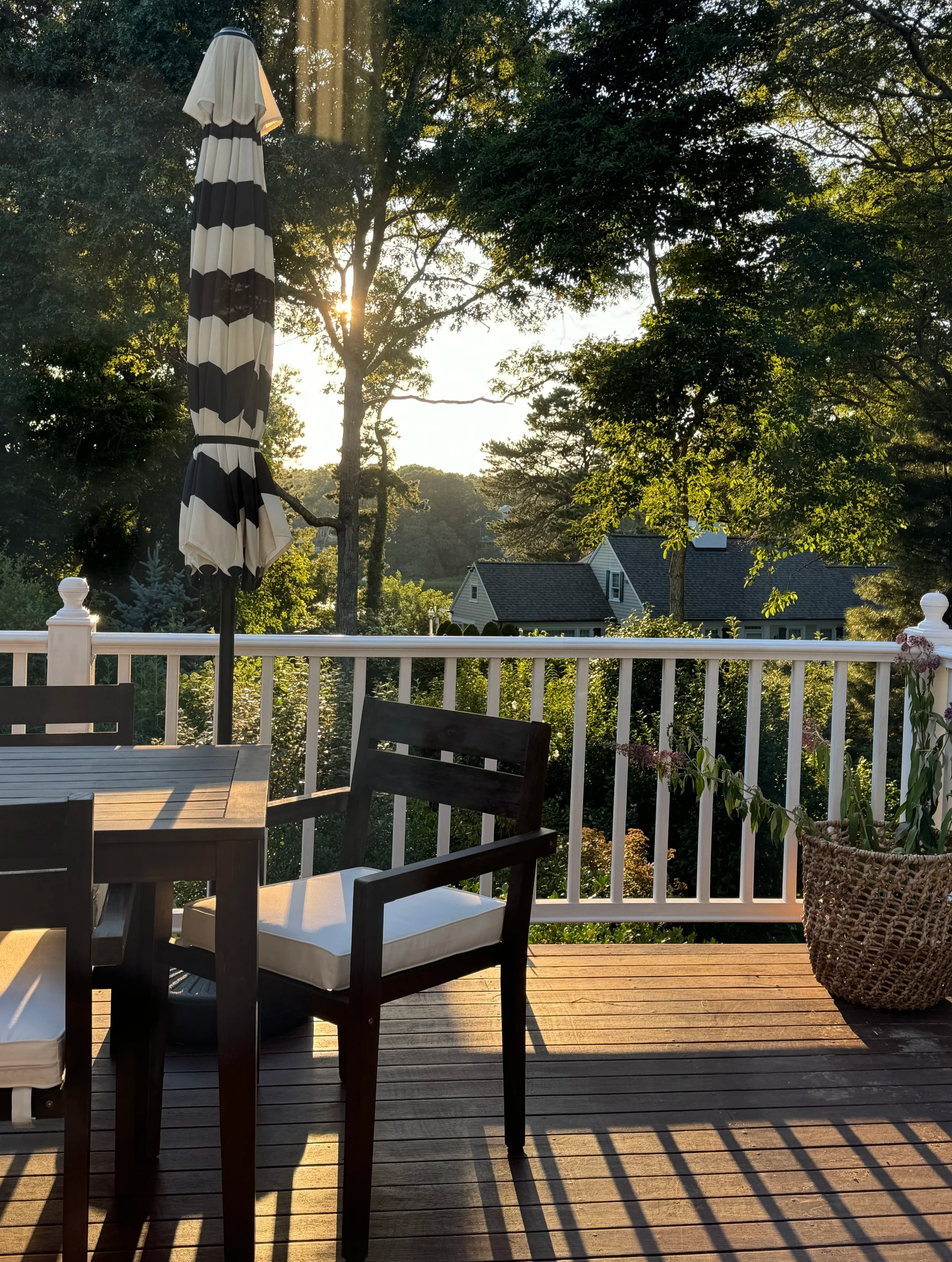 An outdoor wooden deck with a table, chairs, a large striped umbrella, and a basket with plants, overlooking a lush green yard with trees and neighboring houses, illuminated by sunlight.
