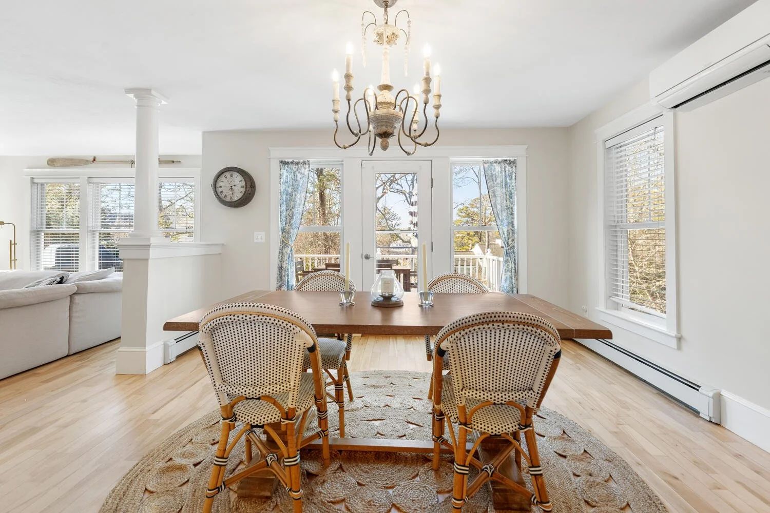 Bright dining room with a large wooden table surrounded by six chairs on a round rug. Large windows and French doors with curtains lead to a deck outside. A chandelier hangs from the ceiling, and there's an air conditioner on the right wall.
