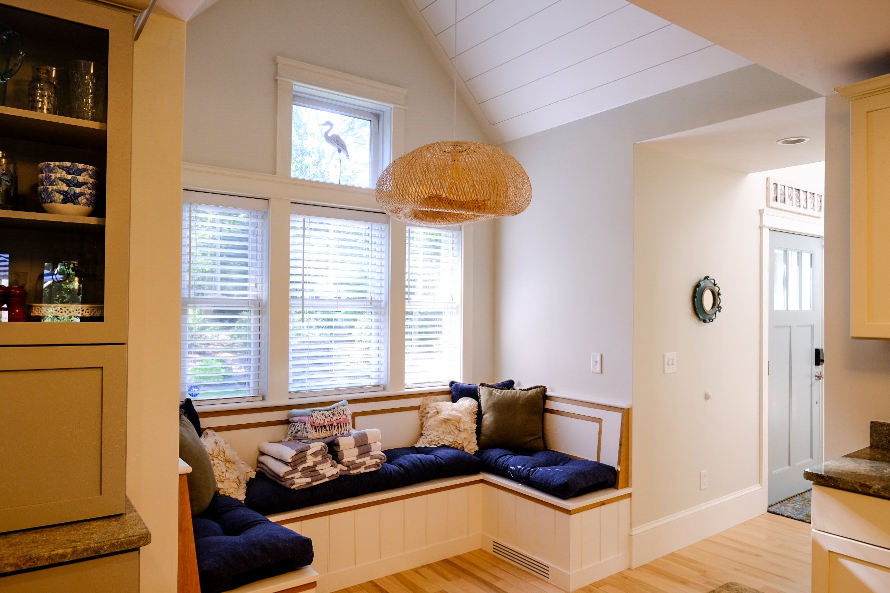 Sunlit kitchen nook with built-in seating, blue cushions, decorative pillows, and stacked linens, next to a window with blinds and a small decorative bird figurine on the window sill.