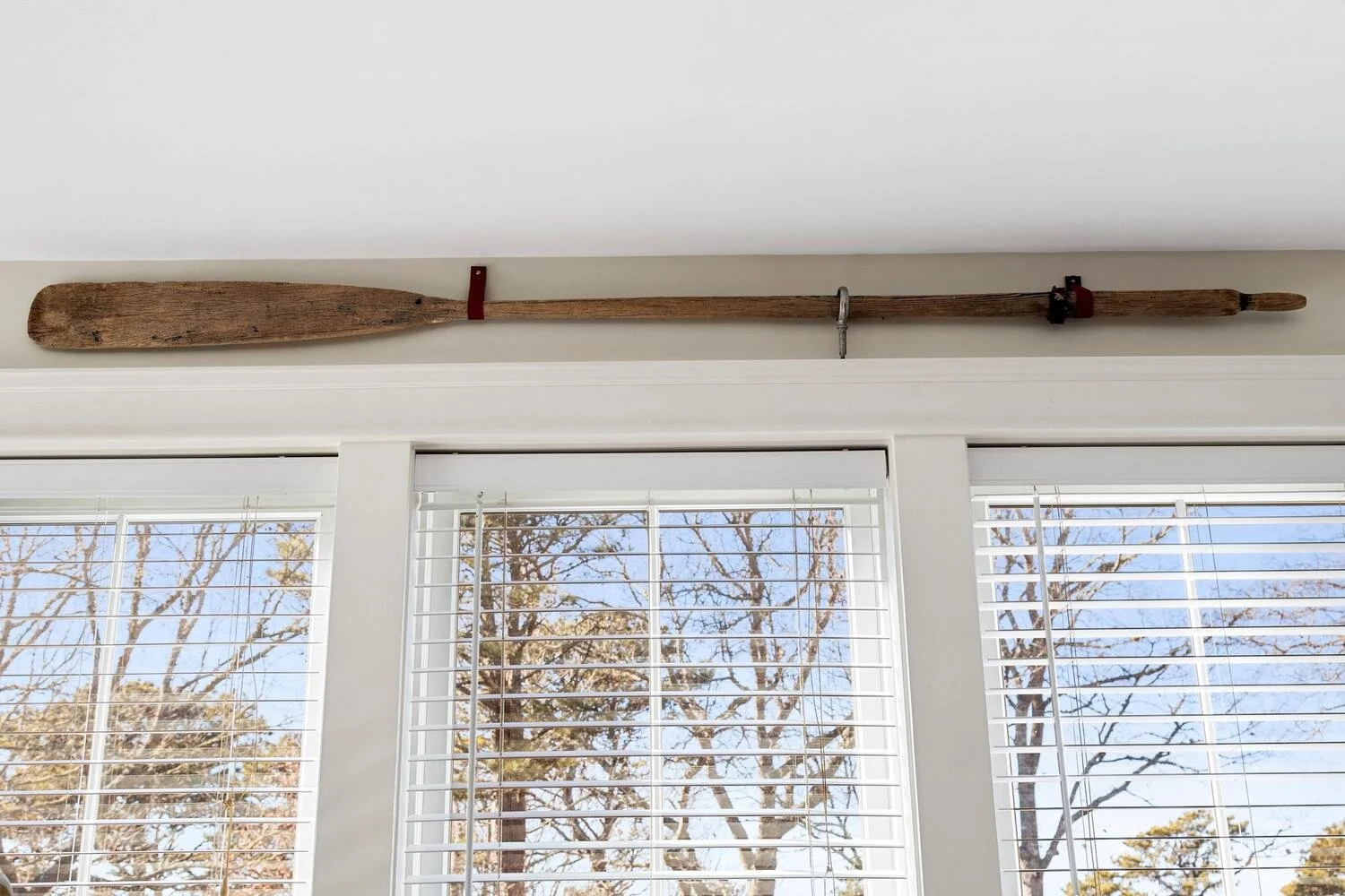 A vintage wooden oar mounted horizontally above a window with white blinds, set against a white wall