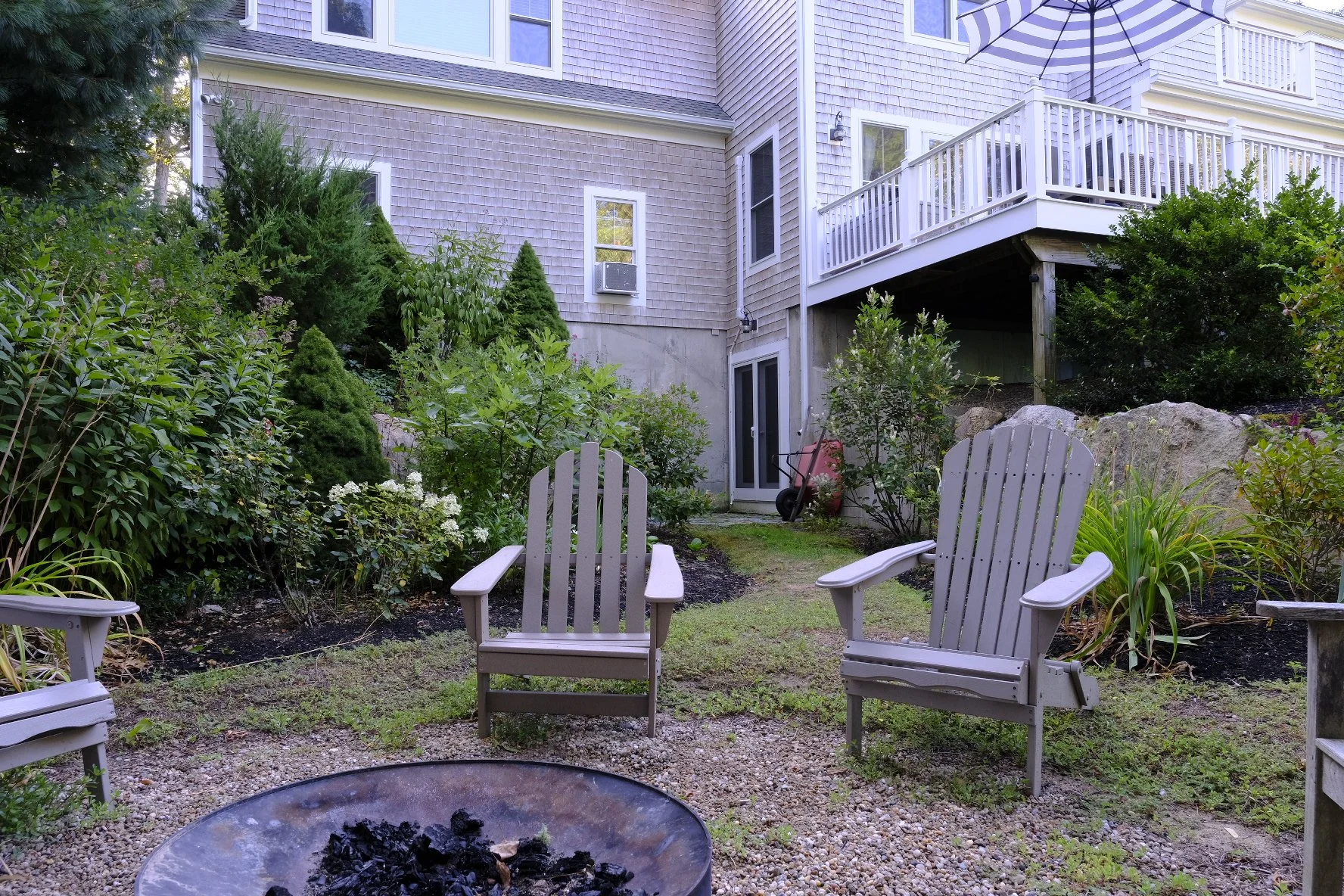Backyard patio with two wooden chairs, a fire pit, lush green bushes, and a house with a balcony and sliding glass door.