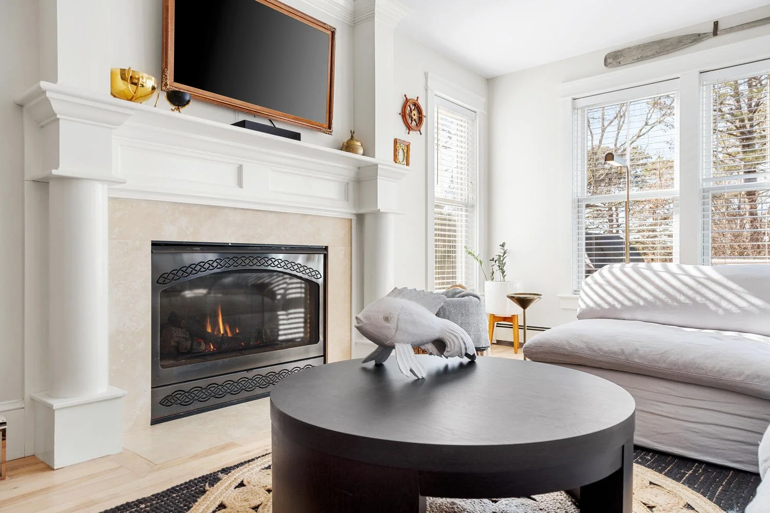 Living room with fireplace, gold and black decorative objects on mantel, flat-screen TV above fireplace, white couch, black coffee table with fish sculpture, large windows with blinds, beige and black rug, sunlight streaming through windows.