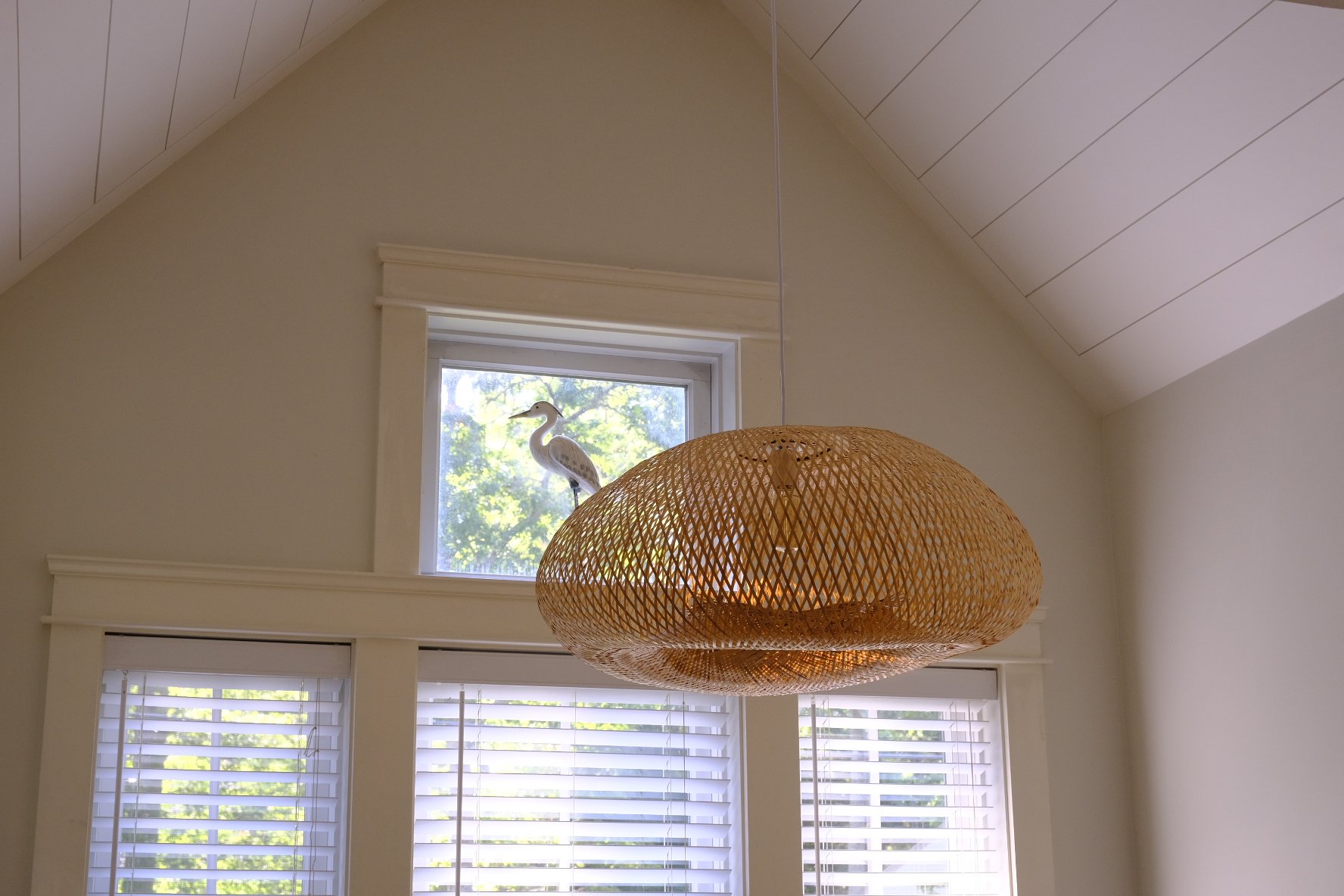 A hanging rattan pendant light fixture in a room with white walls, a sloped ceiling, and windows with white blinds. A decorative bird sculpture is perched on the window ledge outside.