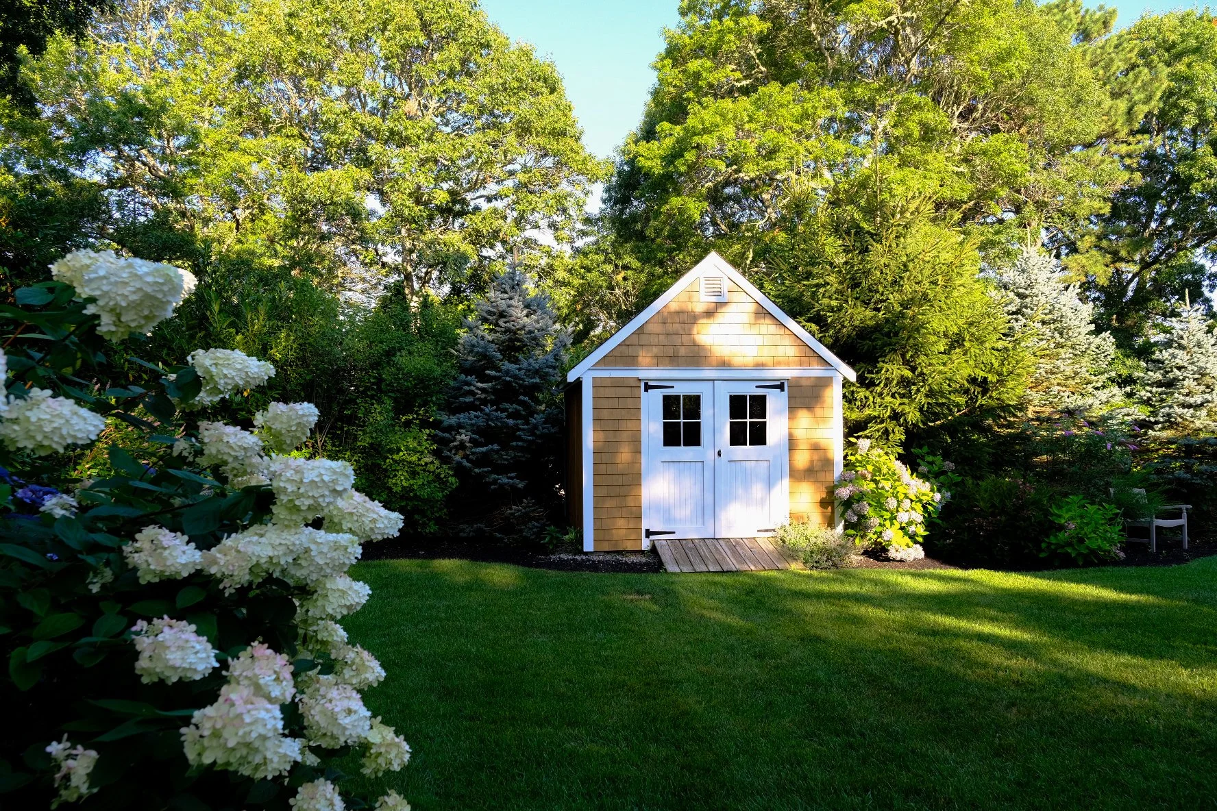 A small wooden shed with white double doors in a lush green backyard, surrounded by flowering bushes and tall trees under a clear sky.
