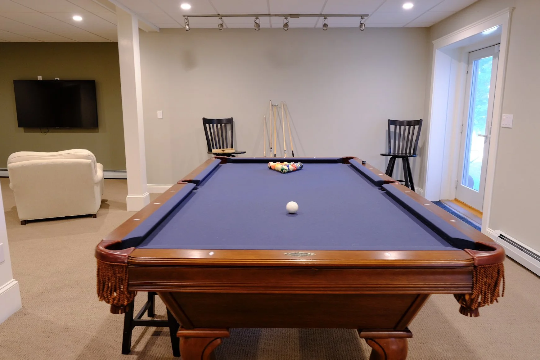 Indoor game room with a pool table at the center, pool balls arranged in a triangle, cue stick on a stand, two chairs, a TV on the wall, a beige armchair, and a door leading outside.