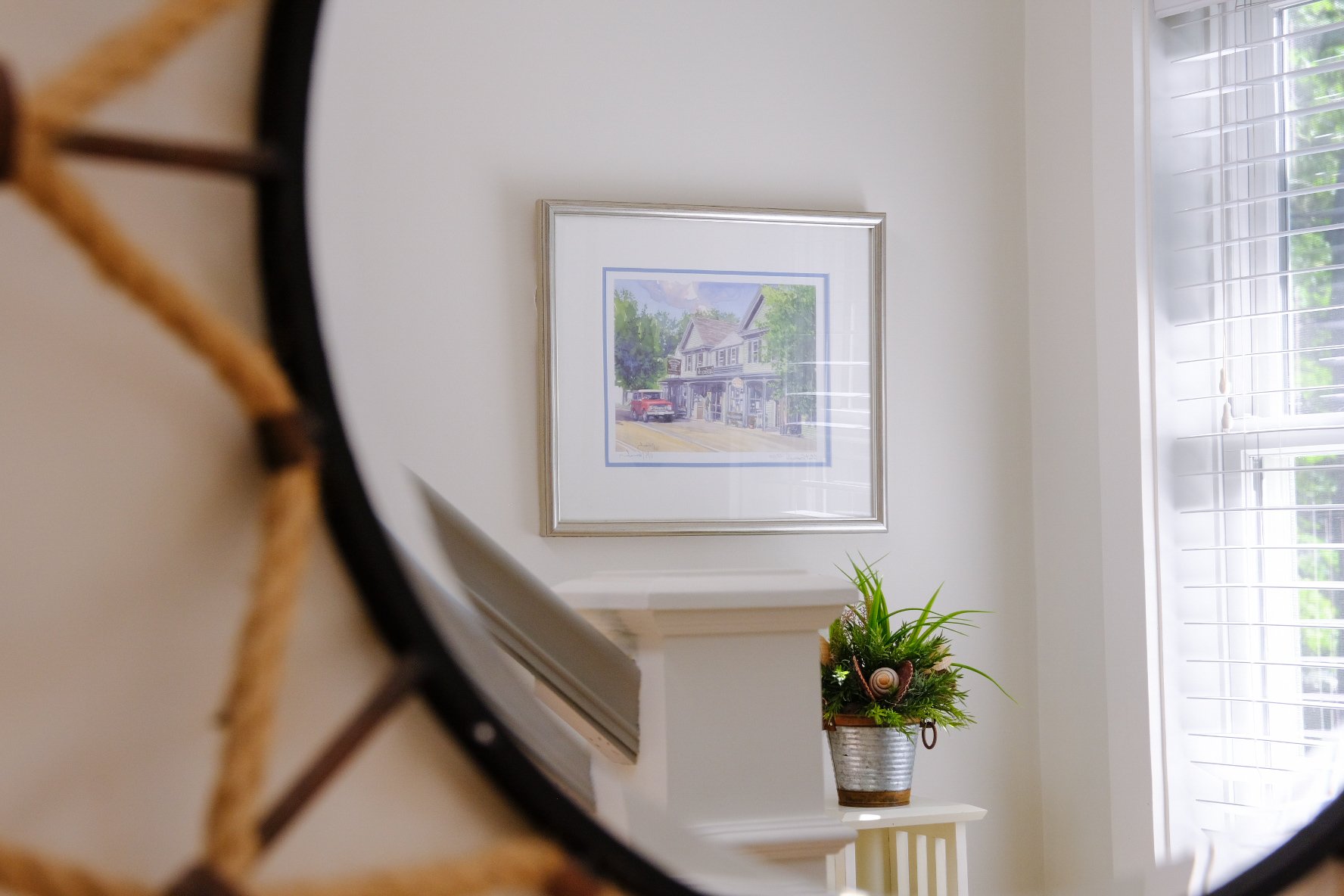 Interior of a home viewed through a mirror, showing a framed picture of a house on the wall, a potted plant on a white table near a window with white shutters.