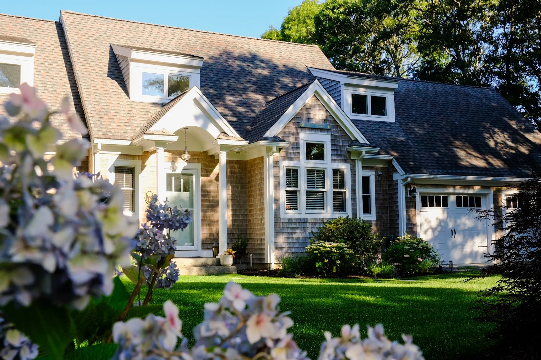 A front view of a large suburban house with a neatly maintained lawn, flowering bushes, and trees, taken during daylight.