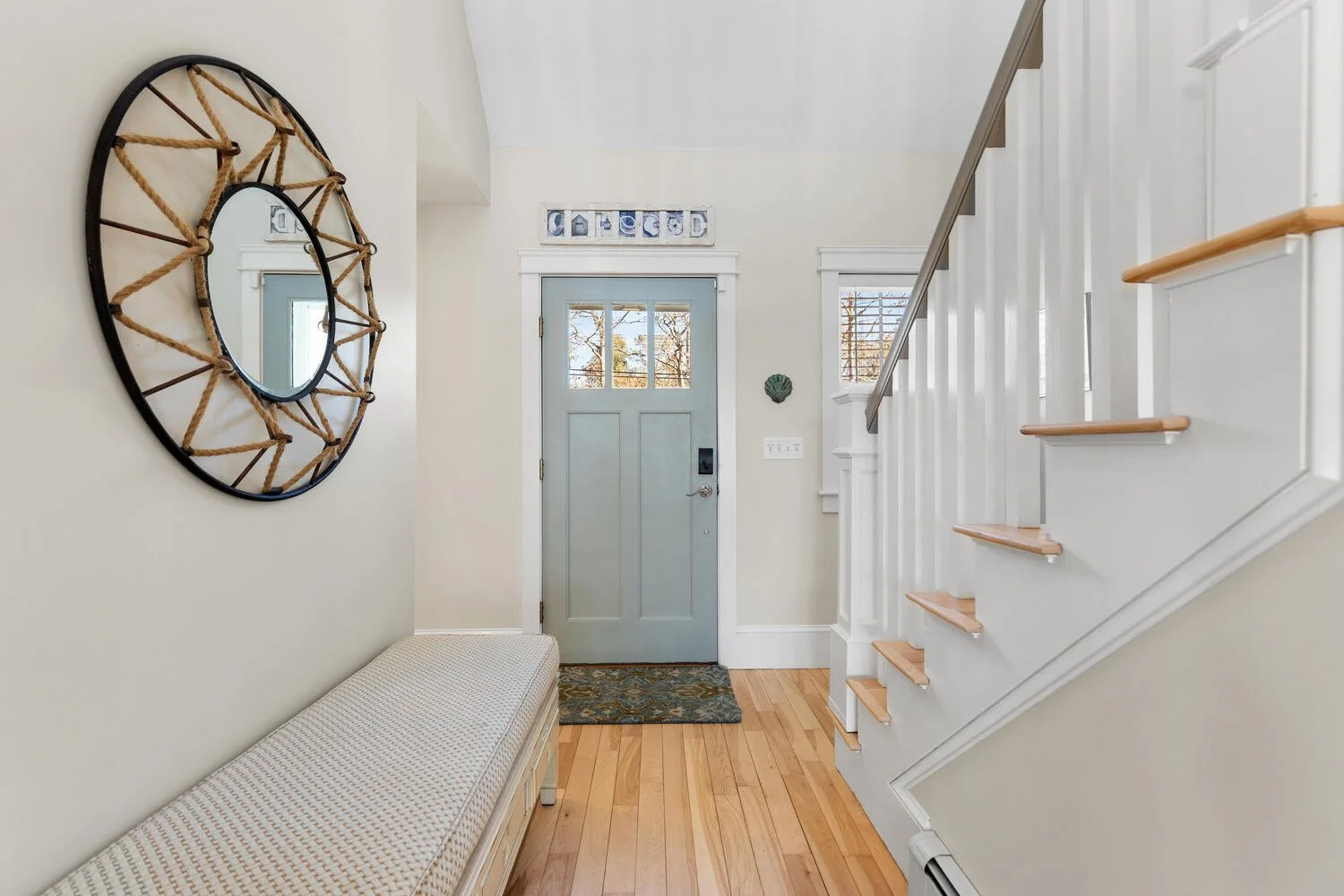 Entryway with light blue front door, white walls, wooden staircase with white risers and light wood treads, wall decor, and a bench along the wall.
