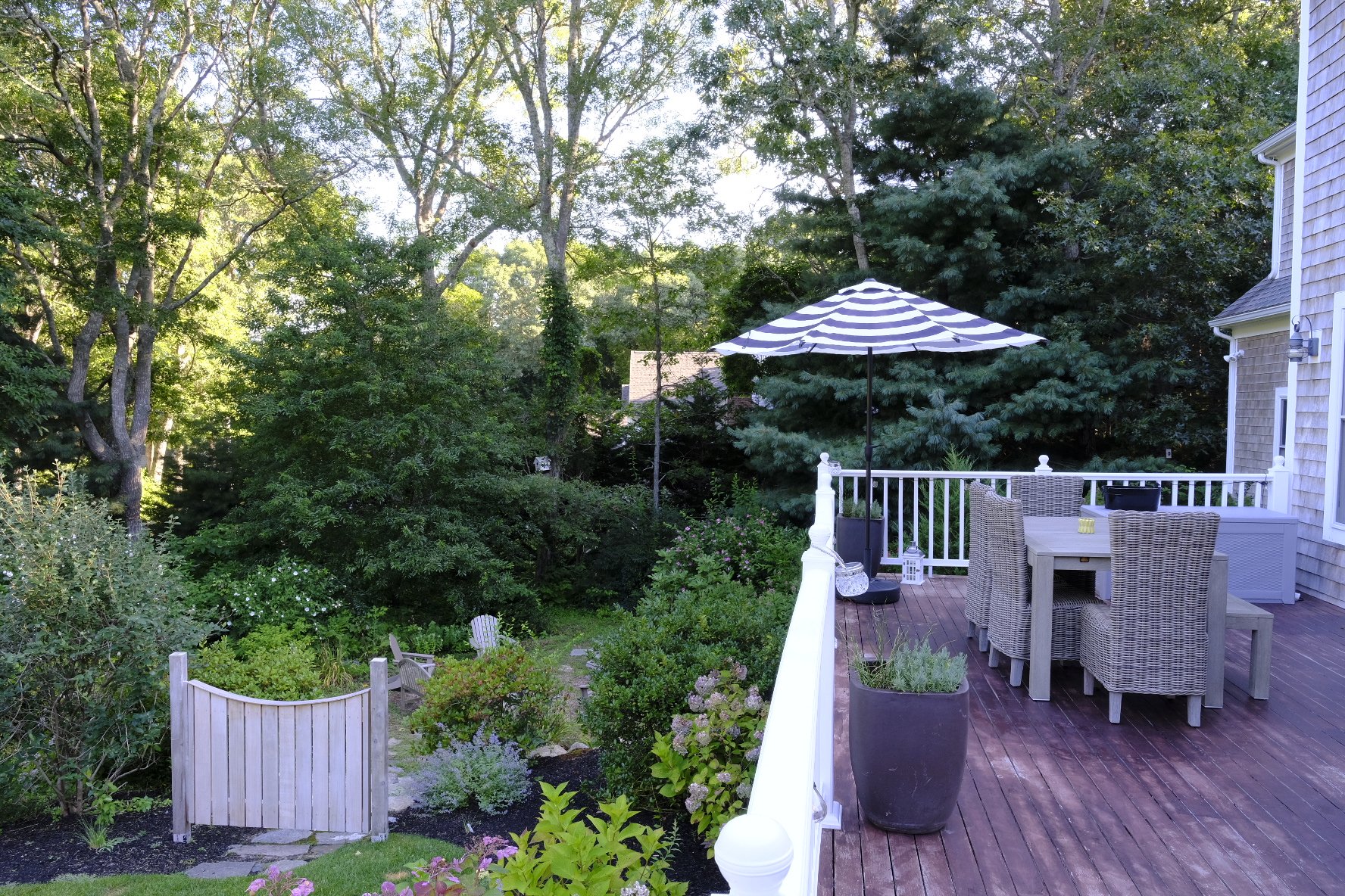 A backyard deck with a striped patio umbrella, wicker chairs, potted plants, a white railing, and lush green trees in the background.