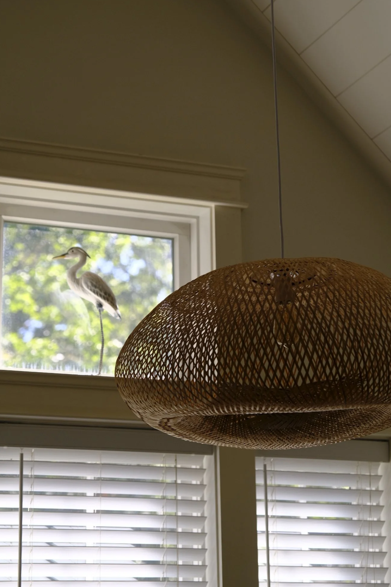 A bird sculpture in a window, with a wicker lampshade hanging inside the room.