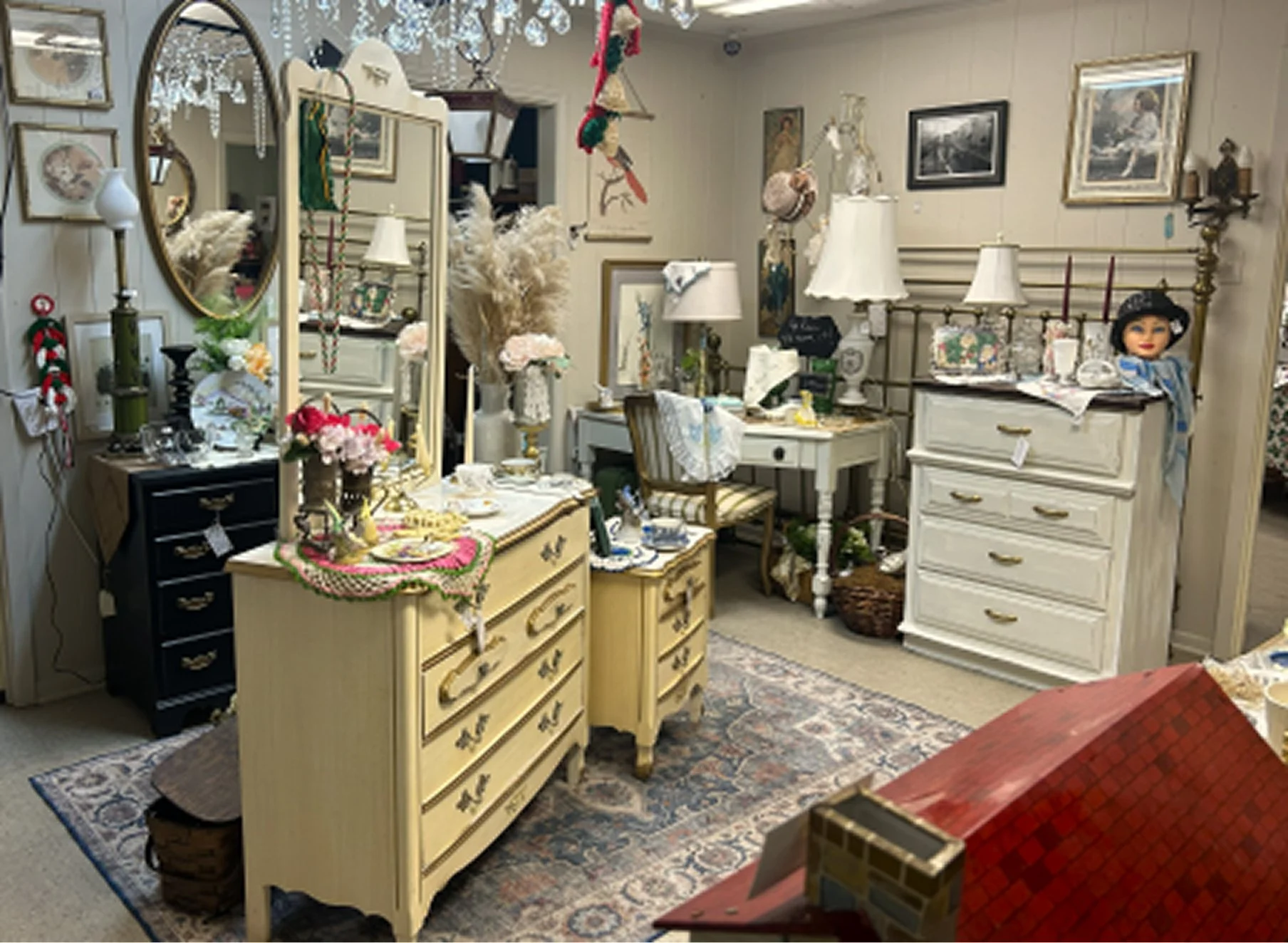 A vintage-style room filled with various antique furniture pieces, decorative lamps, framed art, dolls, and holiday decor including a small doll wearing a witch hat, arranged on dressers, dressers, tables, and walls. A large mirror reflects part of t