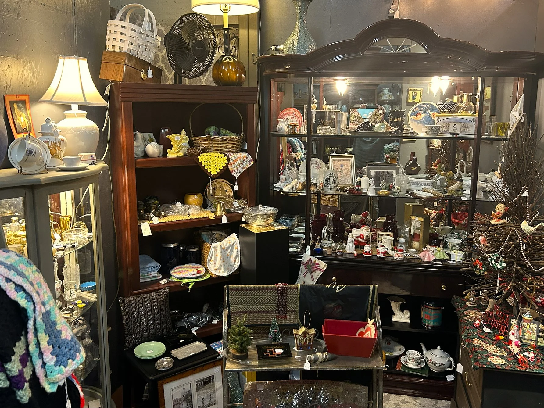 Interior of an antique store with various glassware, ceramics, and decorative items displayed on shelves and tables, including a glass cabinet filled with collectibles and a Christmas decoration on the right side.
