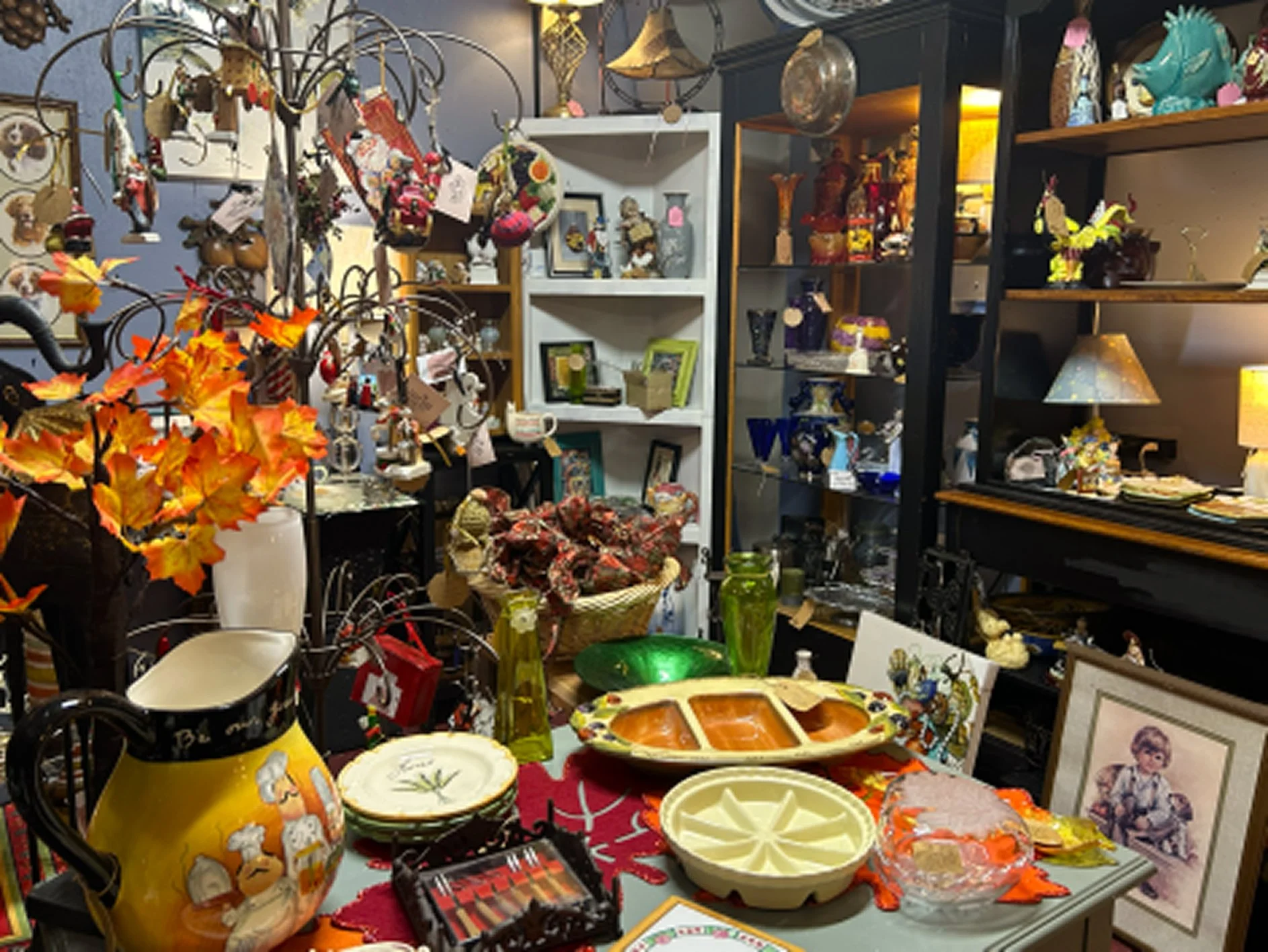 An antique store shelf and table filled with decorative items, ceramics, ceramics, framed pictures, lamps, and fall-themed autumn leaves.