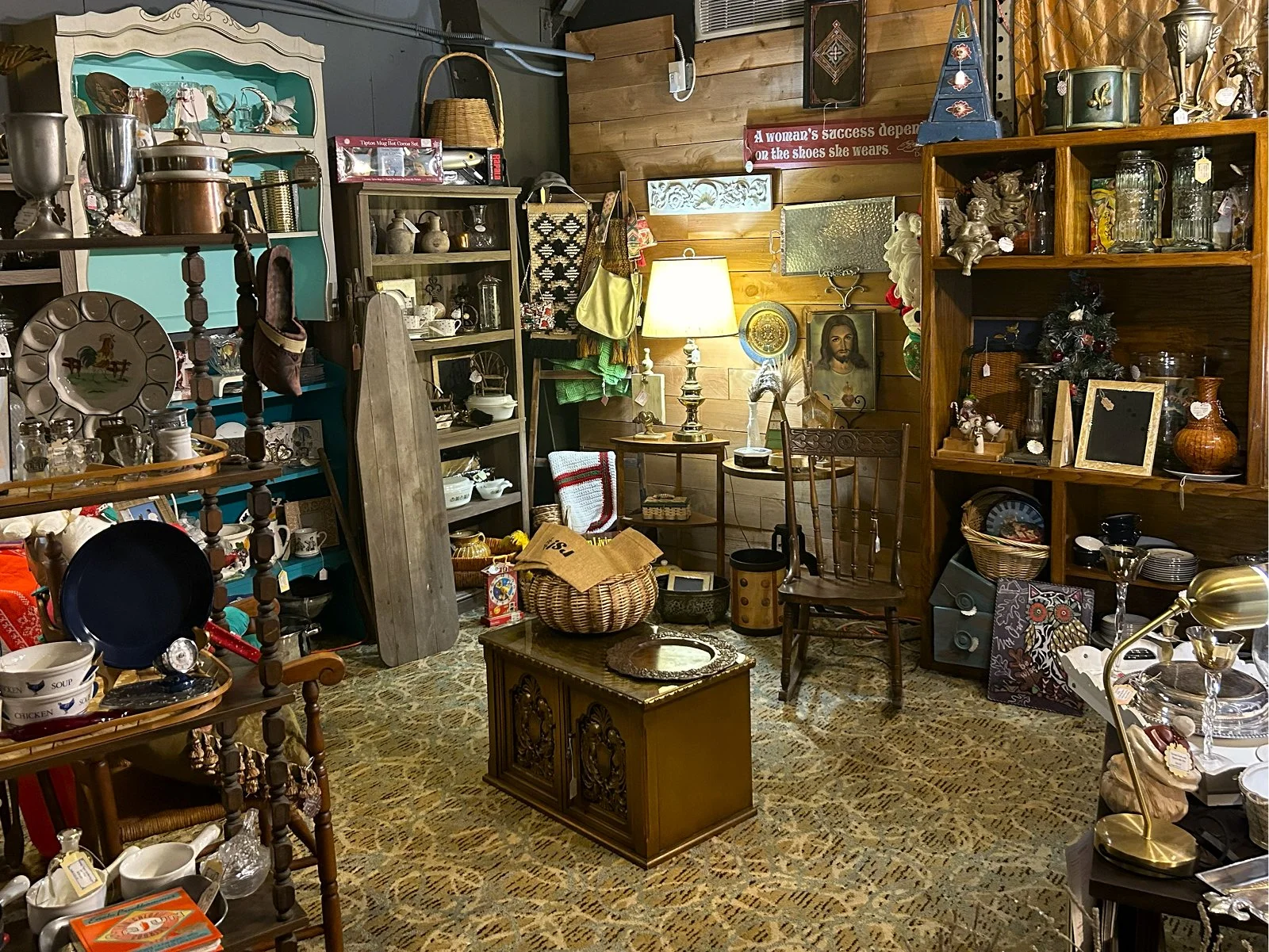 Interior of an antique shop with various shelves and tables filled with vintage household items, decorations, and ceramics. There is a lamp on a side table, a wooden shelf with glassware, and a small central wooden cabinet, all set against a wooden a