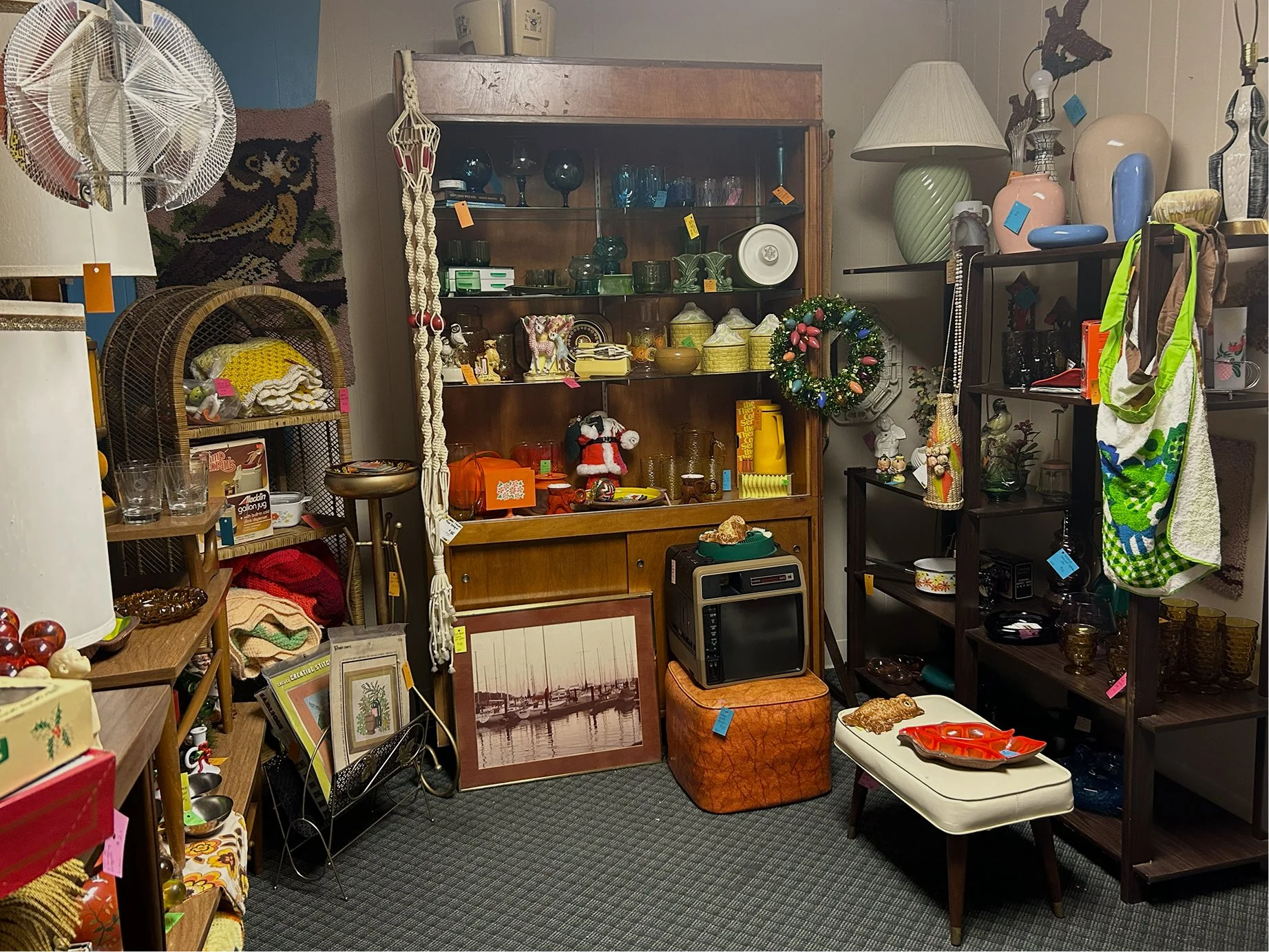A room with various shelving units filled with decorative objects, glassware, and small collectibles. Several lamps, a framed picture of boats, and a small TV are also visible. Items include ceramic vases, a Christmas wreath, and a variety of knickkn