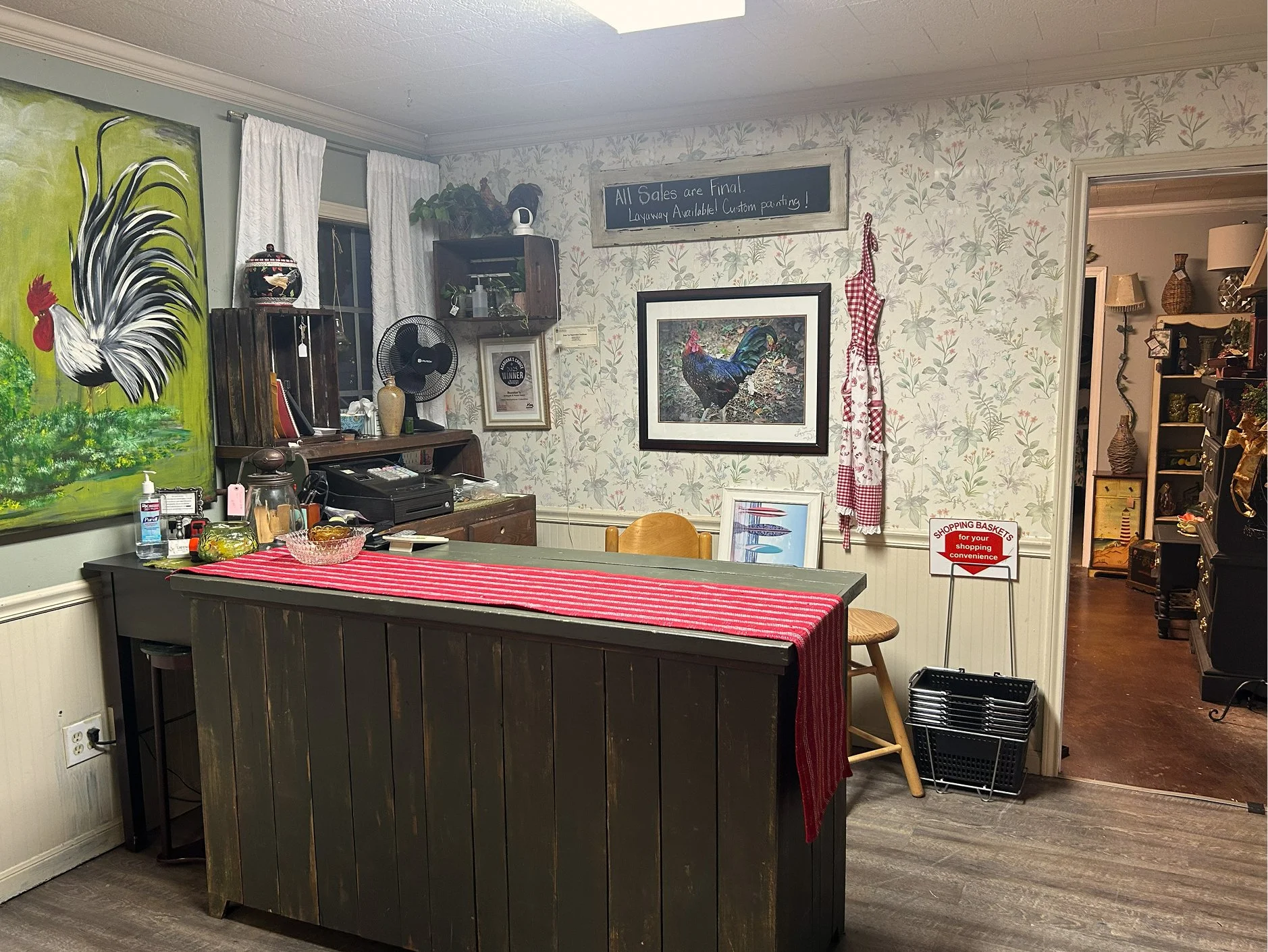 An entryway with a dark wood counter covered with a red and white striped cloth, a painting of a rooster, a chalkboard sign, and various decorations on the wall and shelves. In the background, a doorway reveals a room with shelves and items.