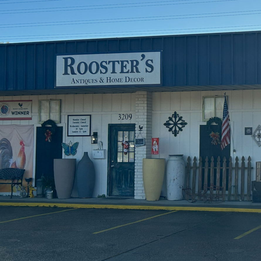 Storefront of Rooster's Antiques & Home Decor with various decorative items outside, including large vases and a fence, and an American flag.