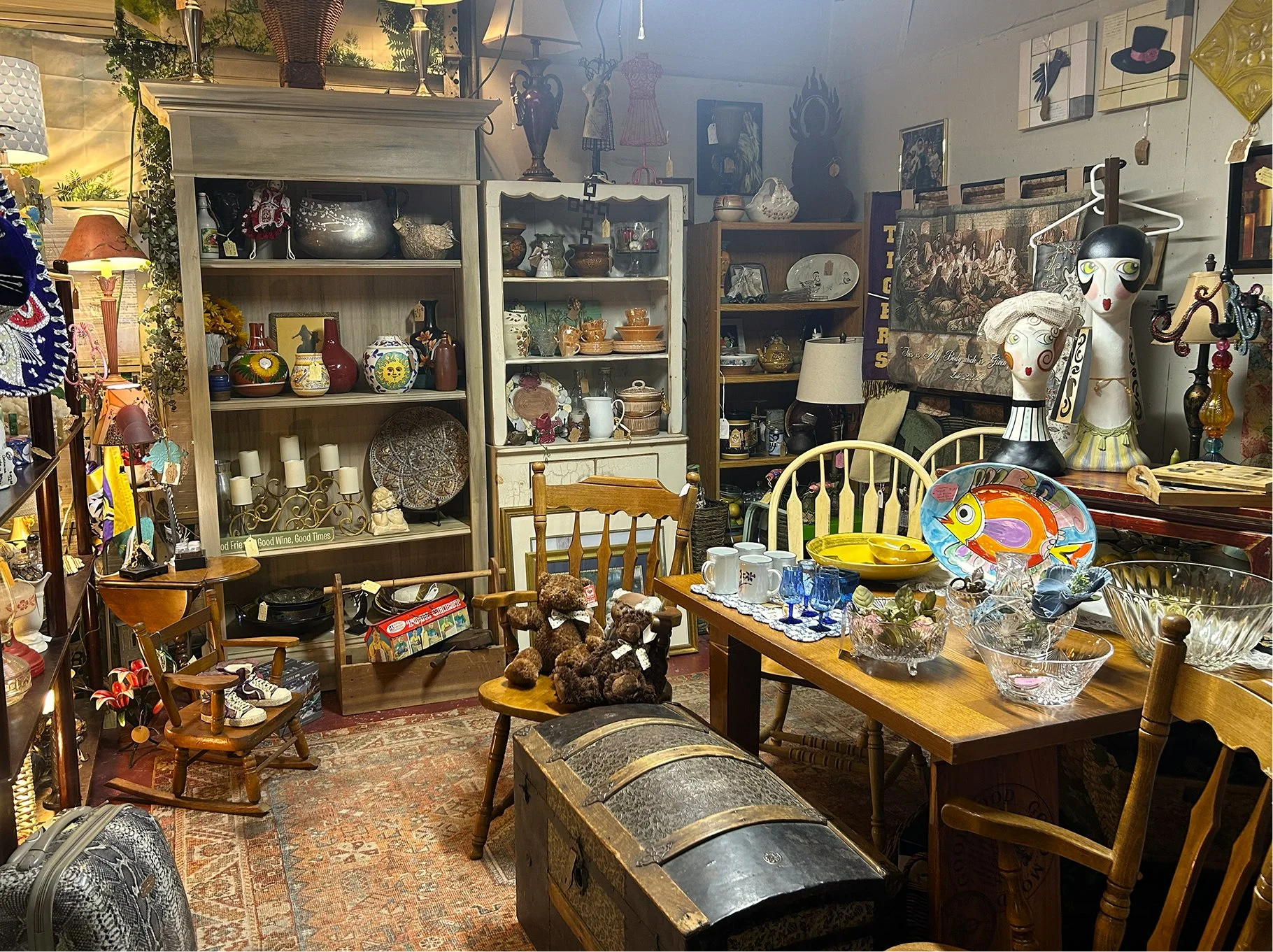 An antique store filled with various vintage items, including shelves of ceramics, glassware, lamps, and artwork. There are wooden chairs, a table with colorful glass and ceramic bowls, teddy bears, and decorative mannequins with stylized faces. The 