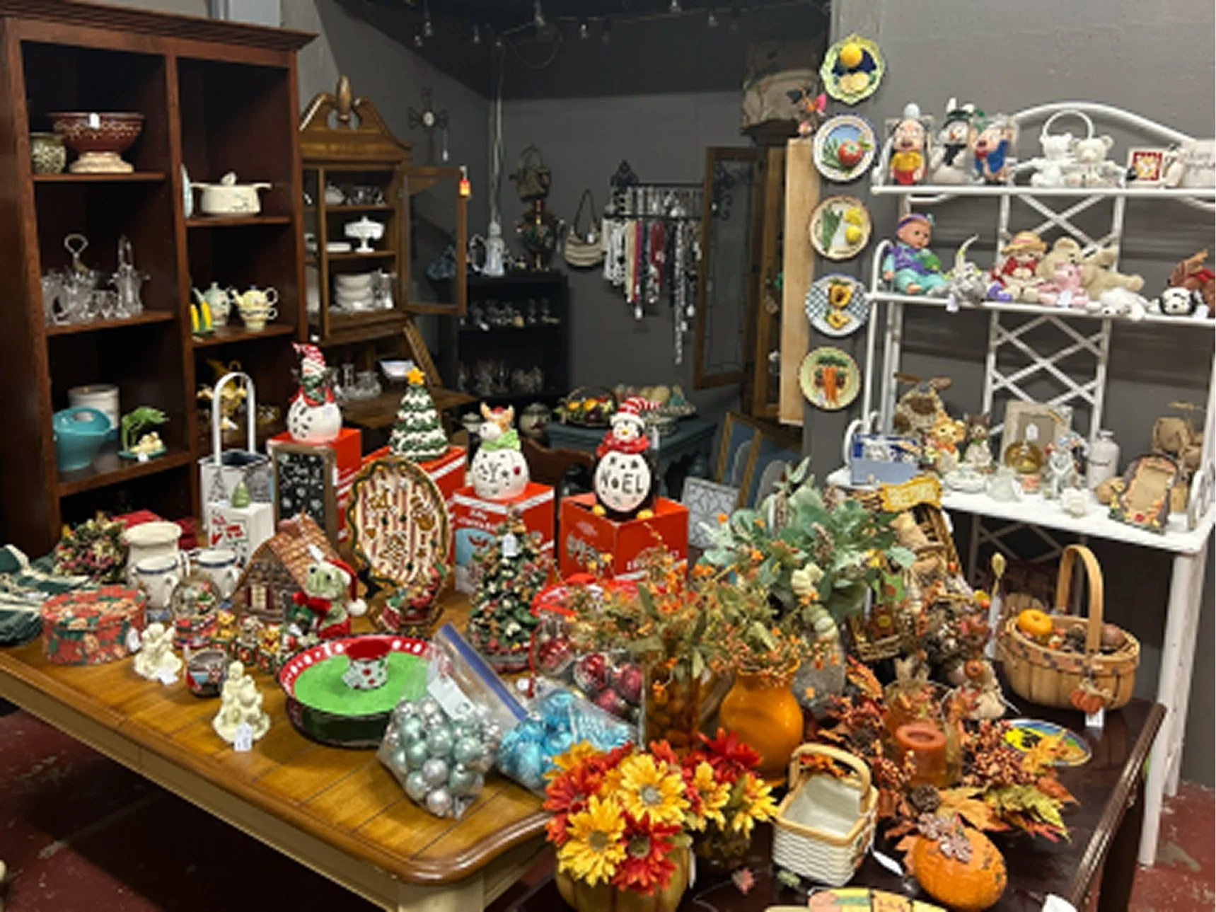 A room with various decorations, figurines, and ornaments on tables, shelves, and display racks, including snowmen, Santa figures, Santa boxes, and artificial fall foliage.