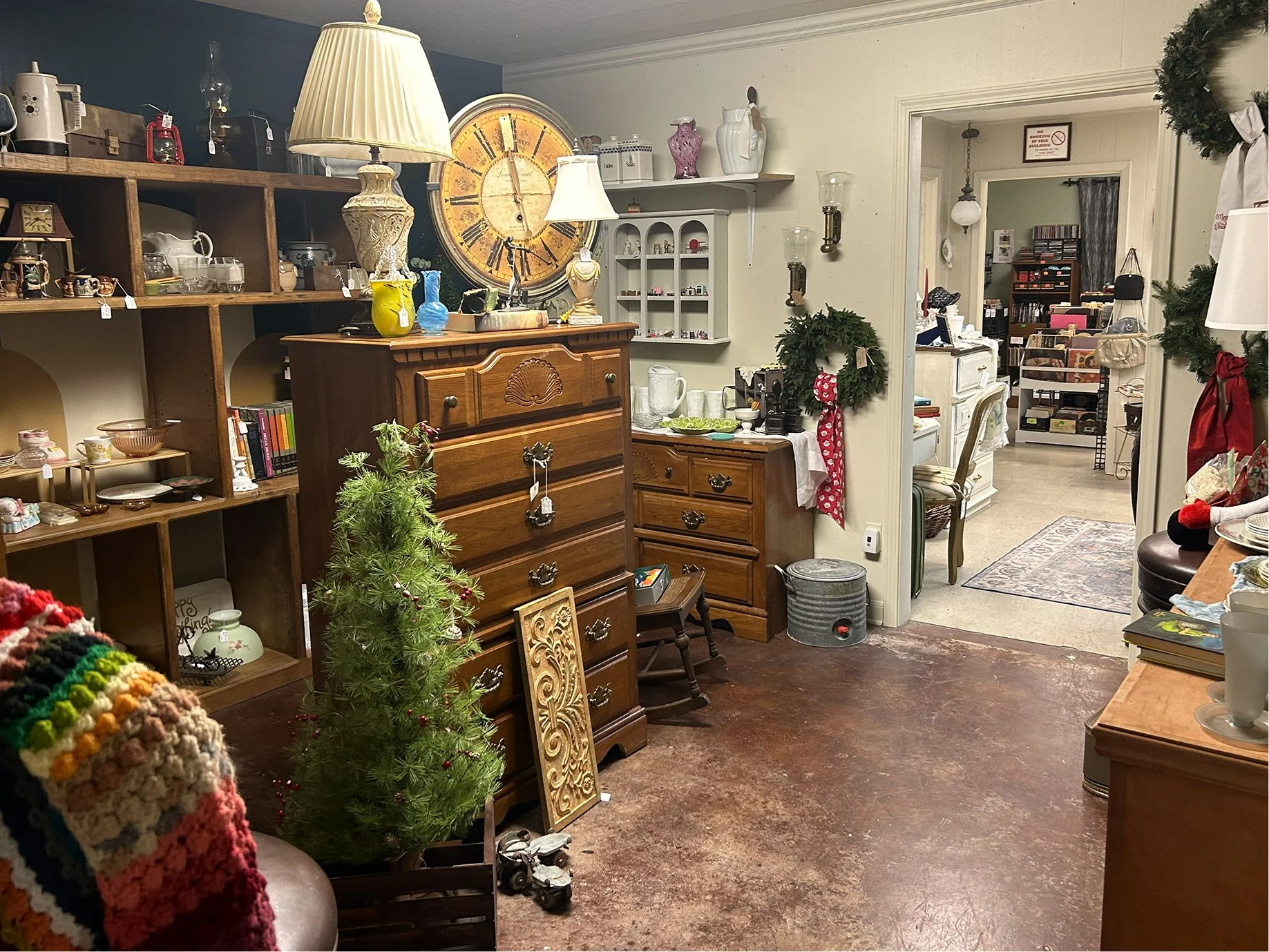 A cozy room decorated with Christmas items, including a small Christmas tree and holiday wreaths, contains various furniture pieces such as a wooden dresser, shelves, and a side table, with decorative lamps, ceramics, and books surrounding the space.