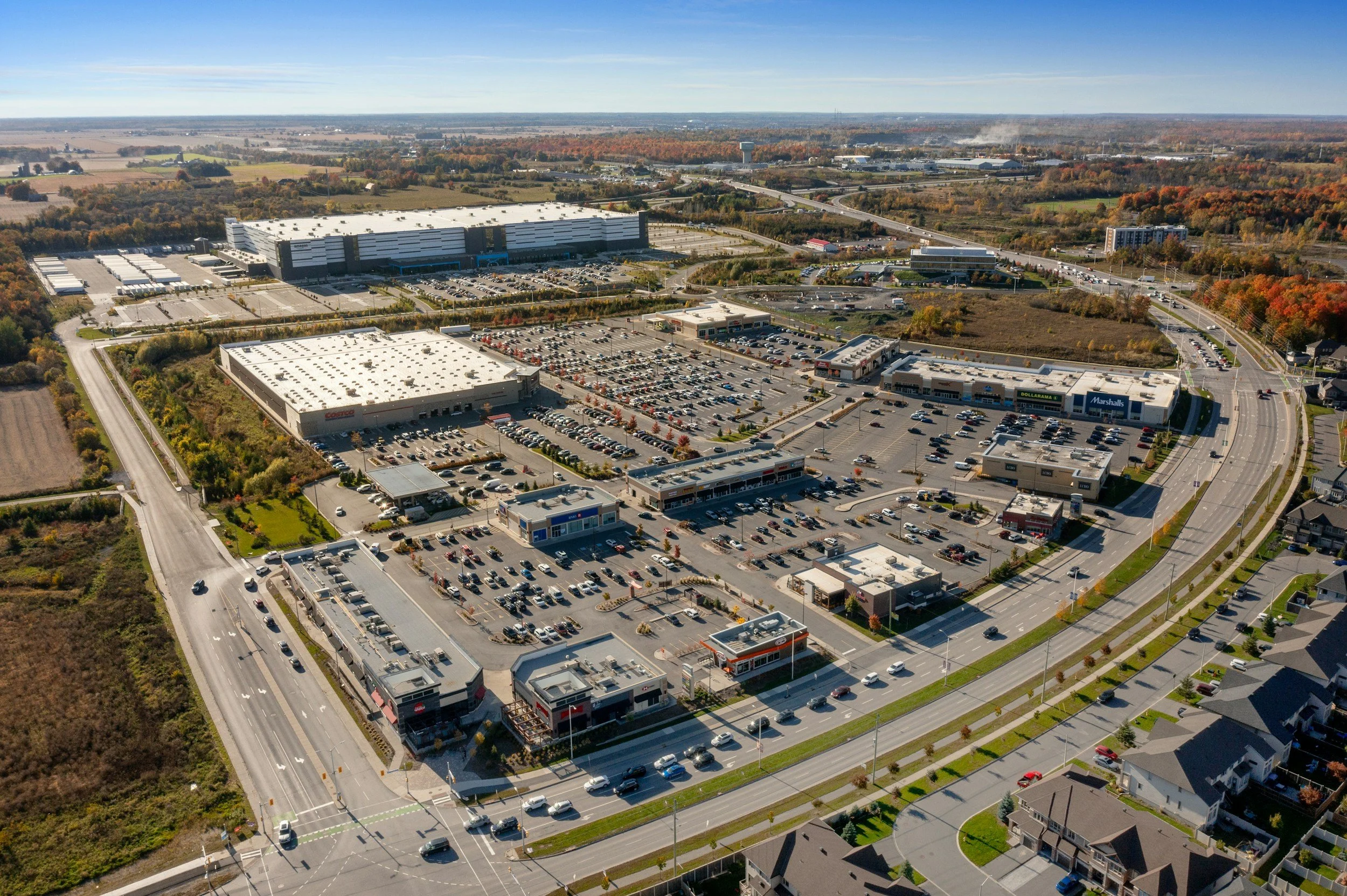 Aerial view of a large shopping plaza with multiple retail stores, parking lots, and surrounding roads in a suburban area during daytime with clear skies.