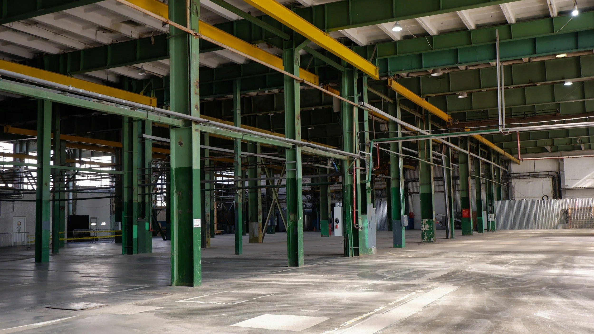 Empty industrial warehouse with green support beams, yellow overhead cranes, and a concrete floor.