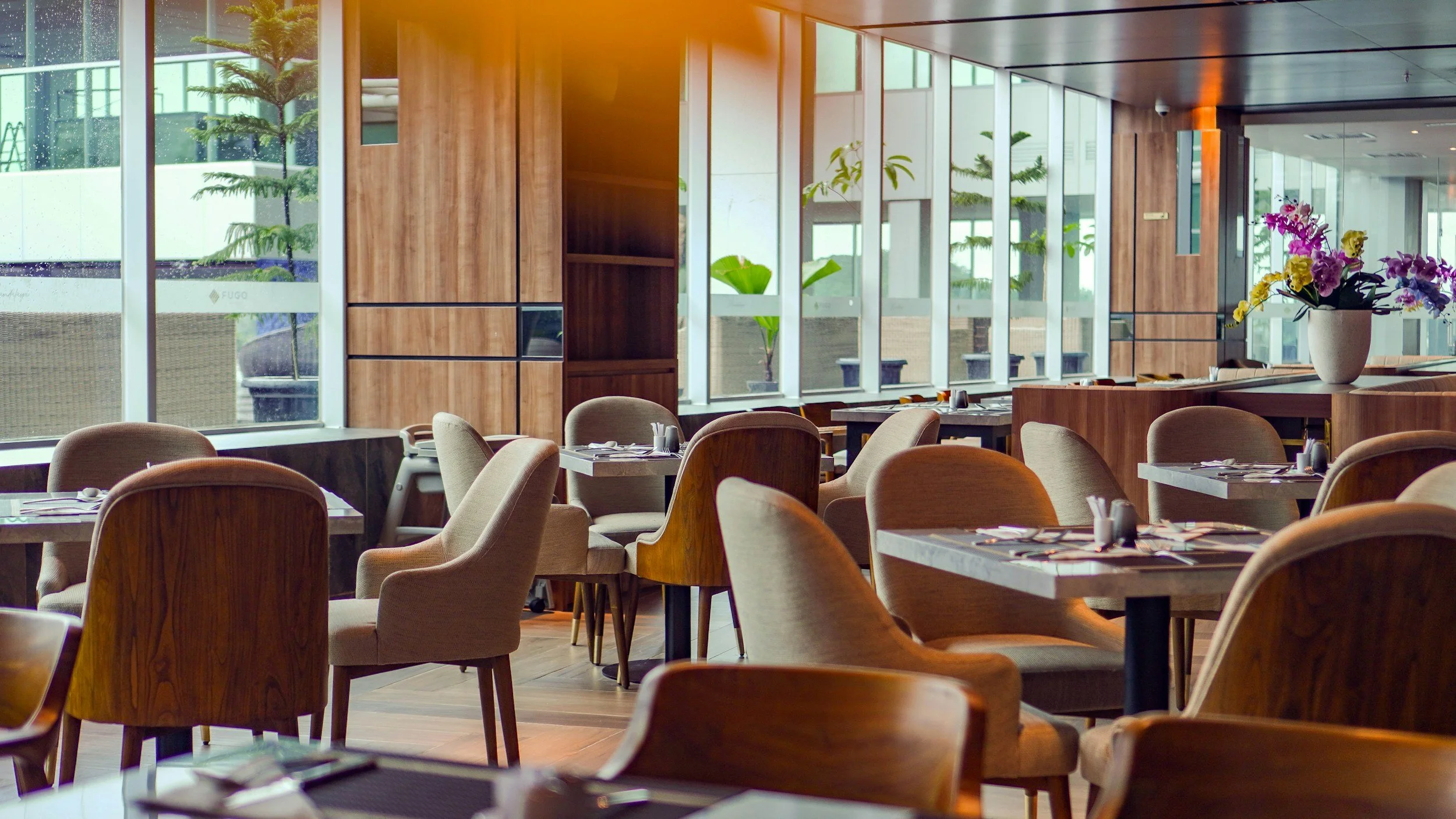 Empty restaurant with wooden chairs and tables, large windows with greenery outside, and a vase with colorful flowers on a counter.