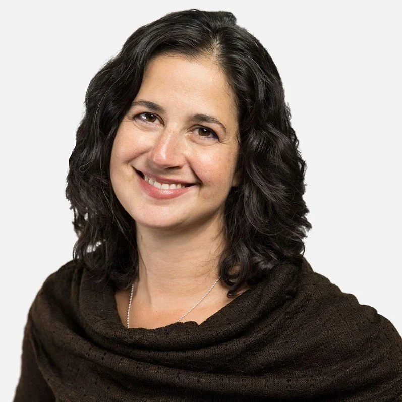 A woman with shoulder-length dark curly hair smiling at the camera, wearing a dark top and a silver necklace against a plain light gray background.