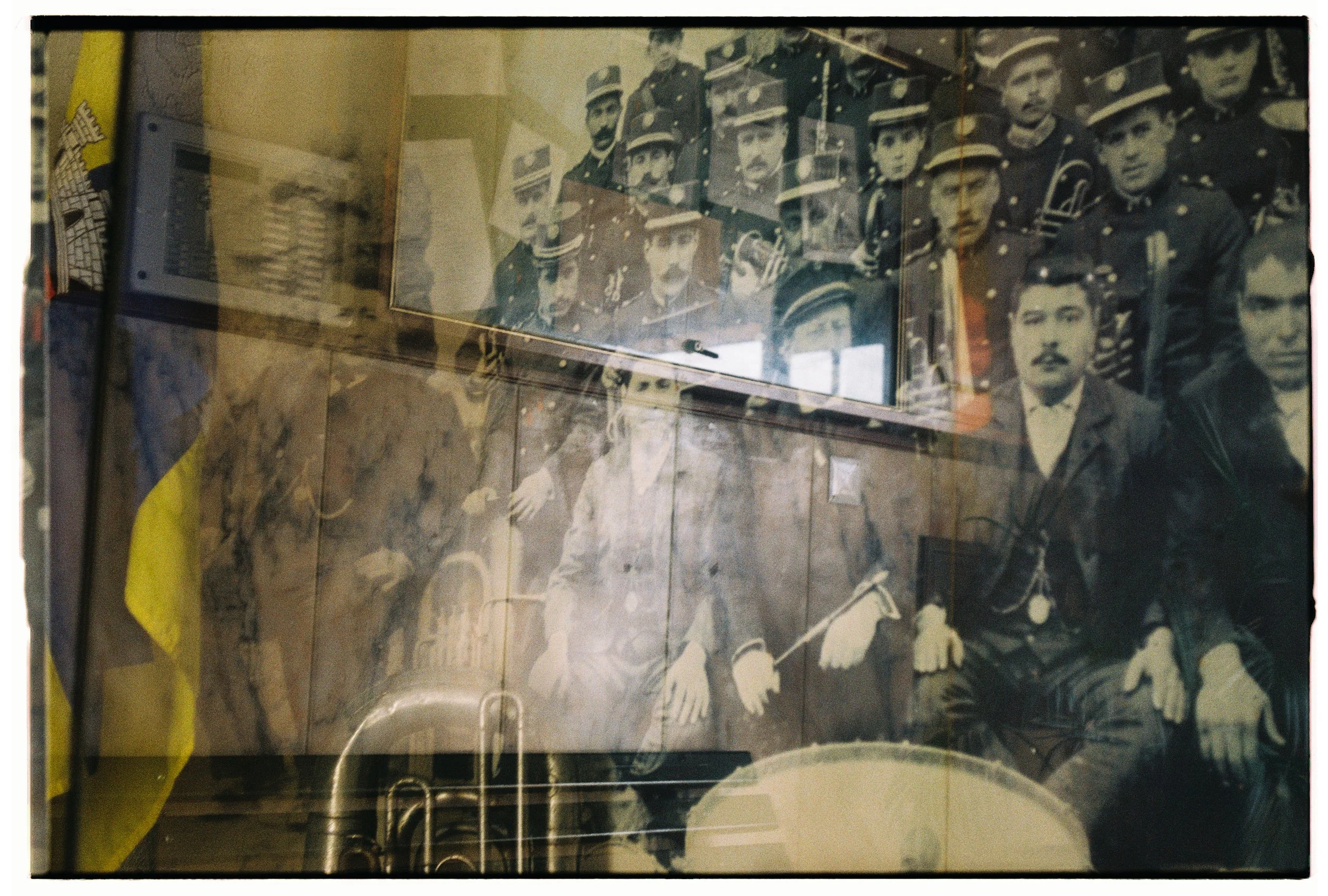 Imagem de um quadro antigo com fotografia de um grupo de homens uniformizados, possivelmente soldados ou policiais, com alguns sentados e outros de pé, exibidos em uma parede de madeira. Reflexo de janela e objetos dentro de uma sala também são visíveis na imagem.