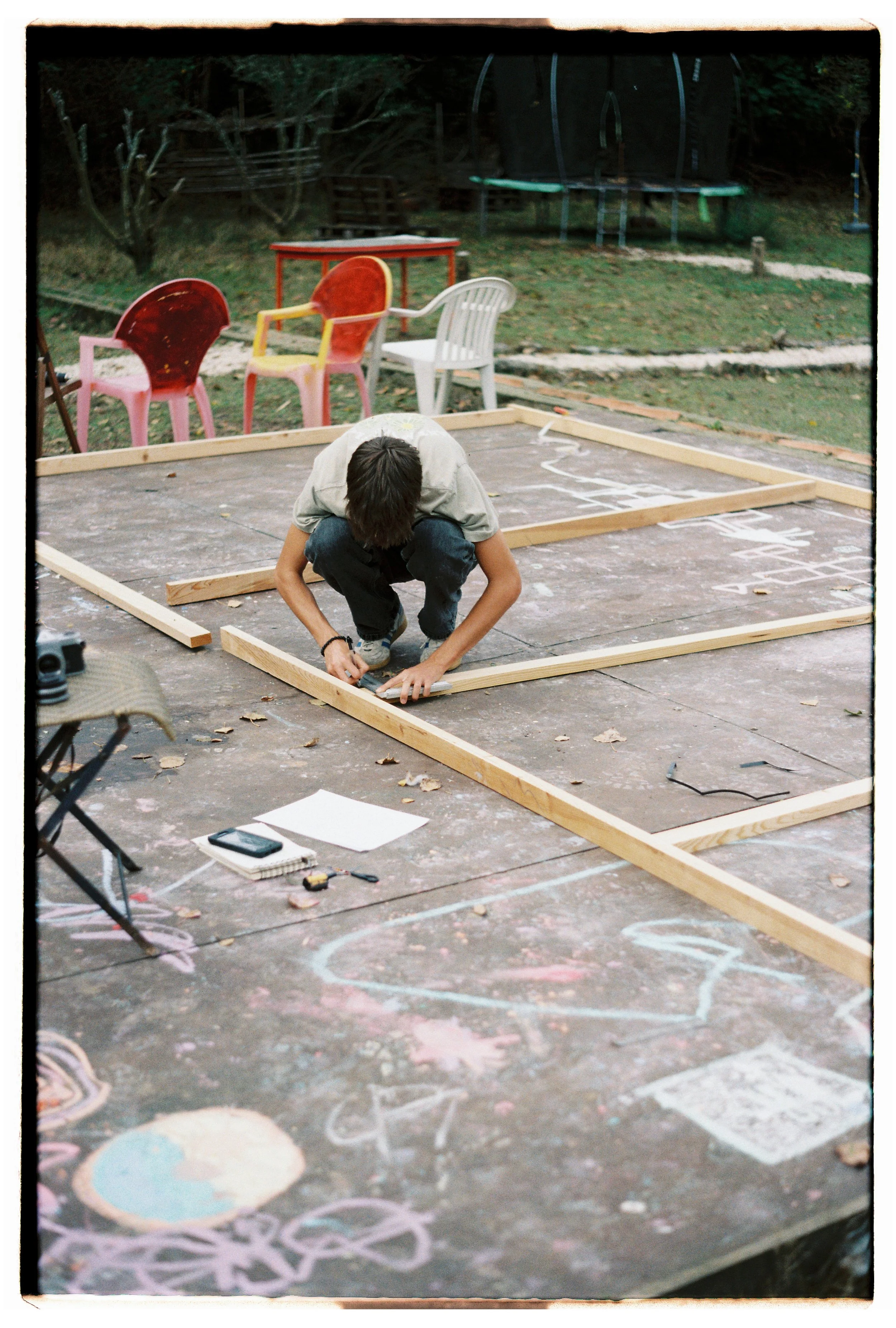 Pessoa montando uma estrutura de madeira em um espaço ao ar livre, com cadeiras coloridas ao fundo e desenhos de giz no chão.