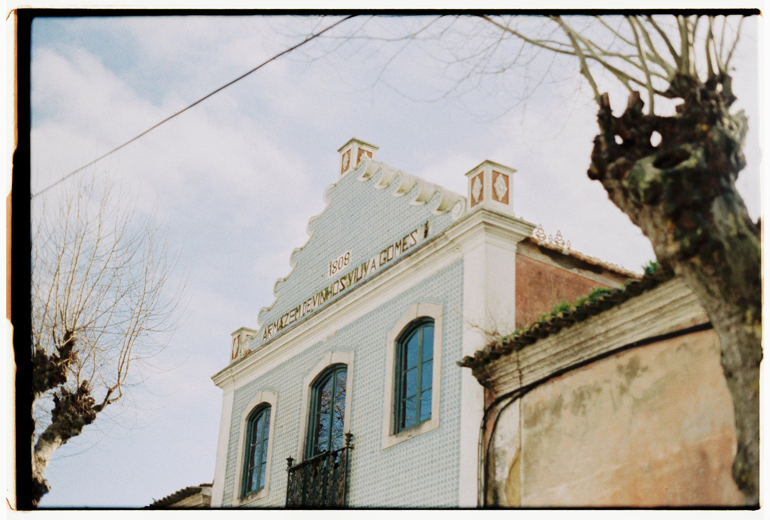 Edifício antigo com fachada decorada, janelas grandes e uma inscrição no topo que diz 'ARMAZÉM DE VEÍCULOS DA GOMES 1808'. Árvores sem folhas ao redor e céu nublado.