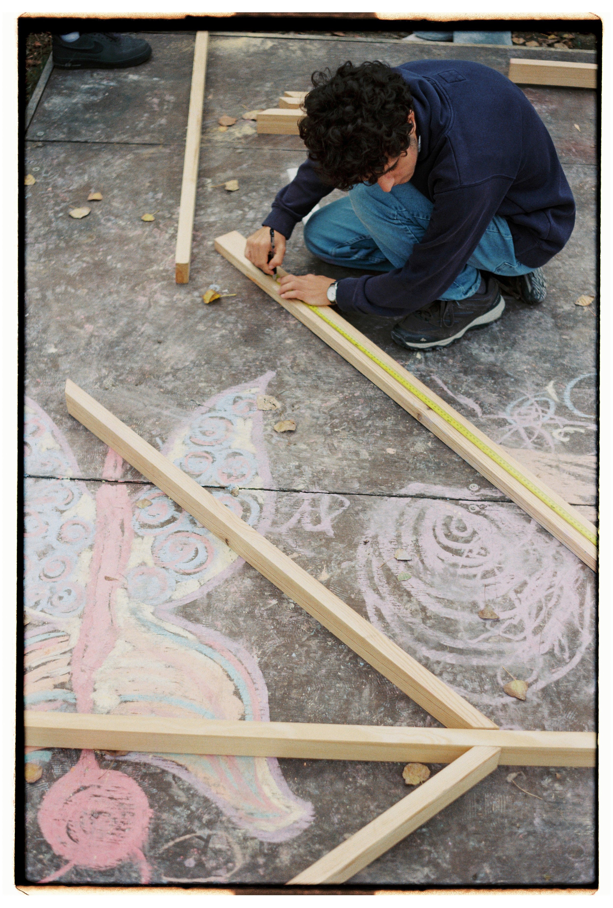 Pessoa jovem fazendo marcação em uma tábua de madeira com régua e lápis ao lado de desenhos coloridos feitos com giz em um chão de concreto.