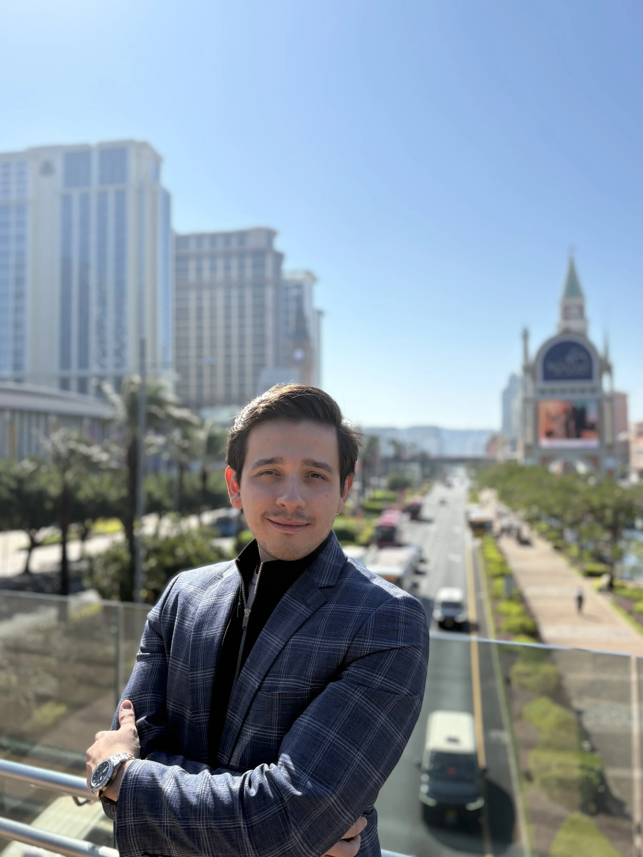 A man in a suit with crossed arms smiling outdoors with city buildings in the background.