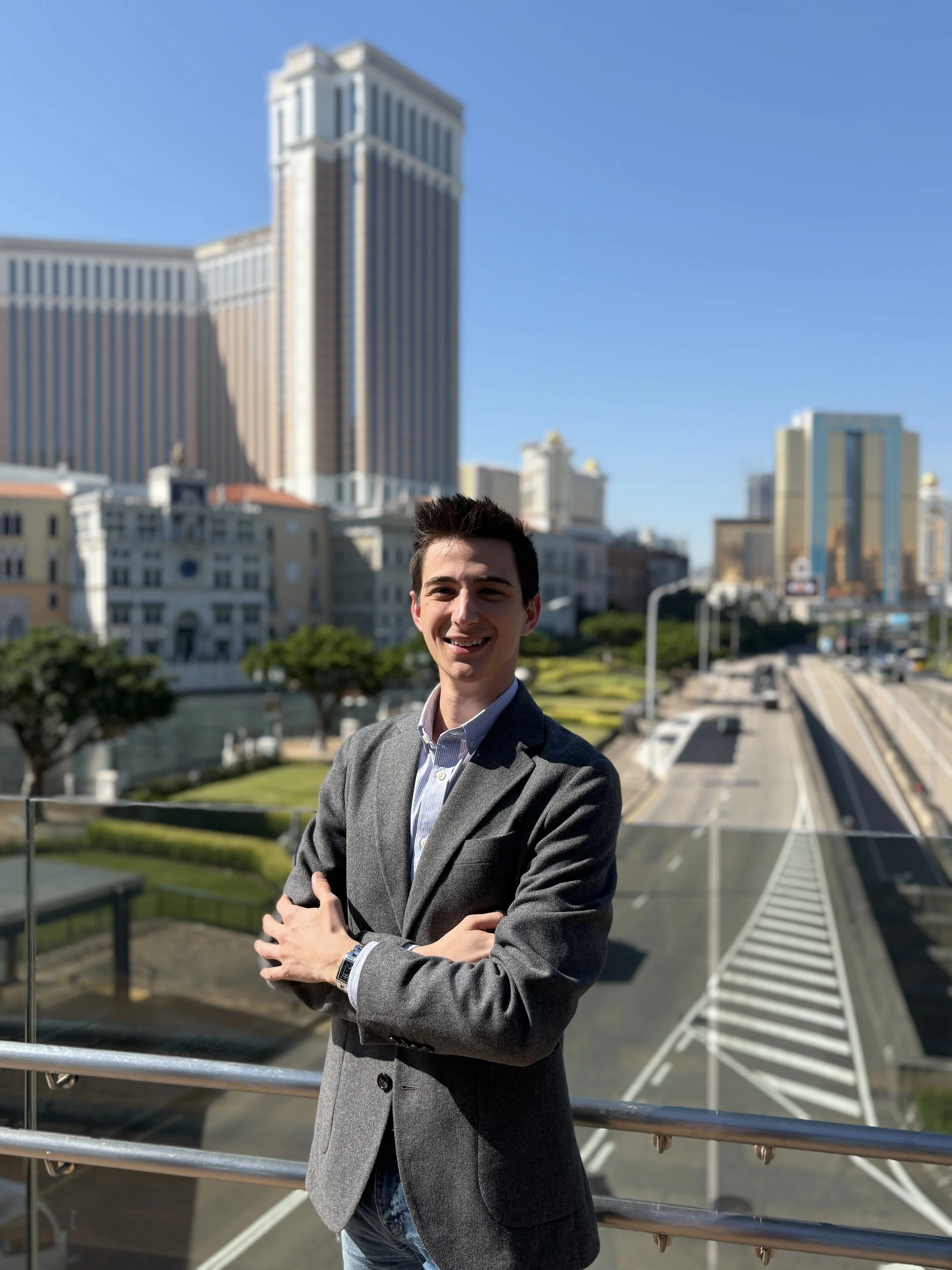 A young man in a gray blazer and shirt standing with arms crossed, smiling outdoors in front of a cityscape with tall buildings and a clear blue sky.
