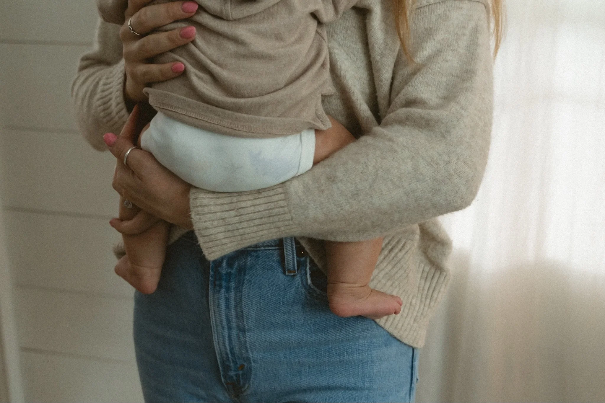 A person holding a baby, focusing on the child's back and the person's hands. The person is wearing a beige sweater, and the baby is dressed in a beige top and white diaper. The person's hands have rings and pink nail polish.