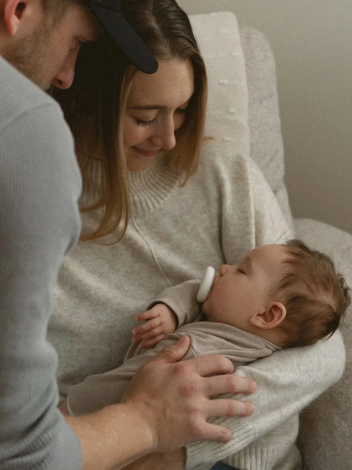 A woman holding a sleeping baby with a pacifier in its mouth, while a man gently touches the baby's arm.