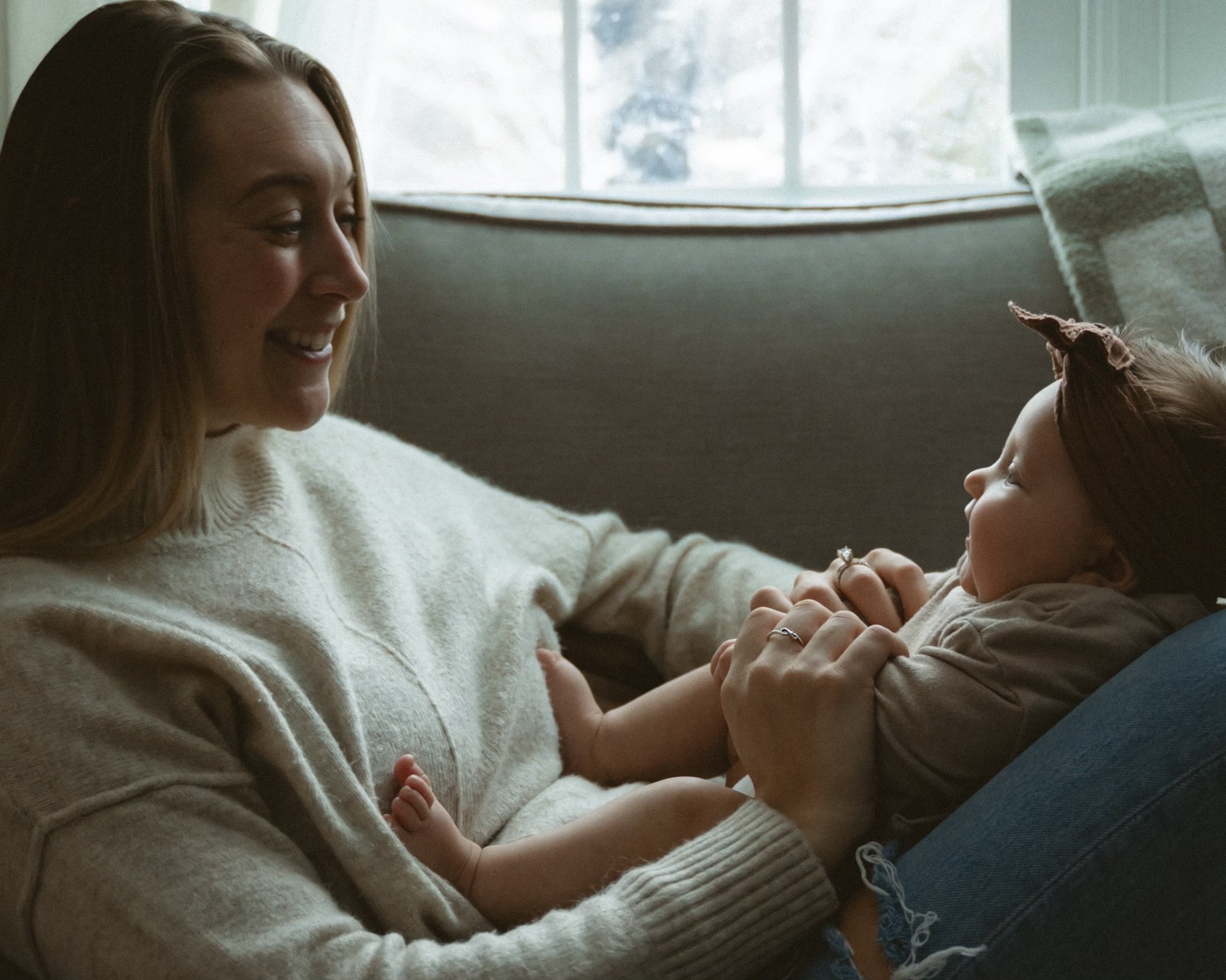 A woman and a young girl sitting on a couch, laughing and looking at each other, with natural light coming through a window in the background.