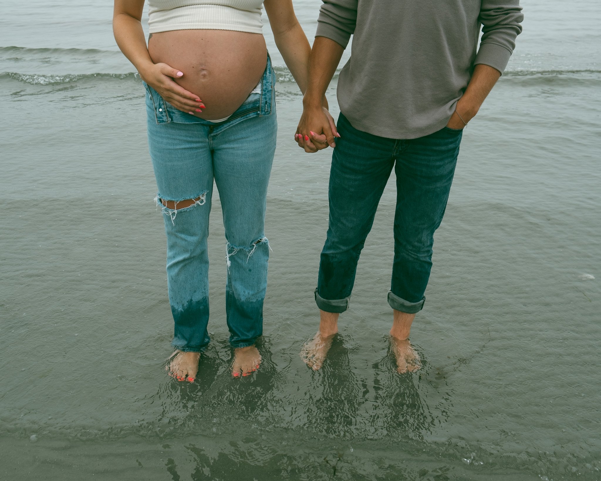 A pregnant woman and her partner holding hands while standing in the shallow ocean water, with their feet submerged and waves in the background.