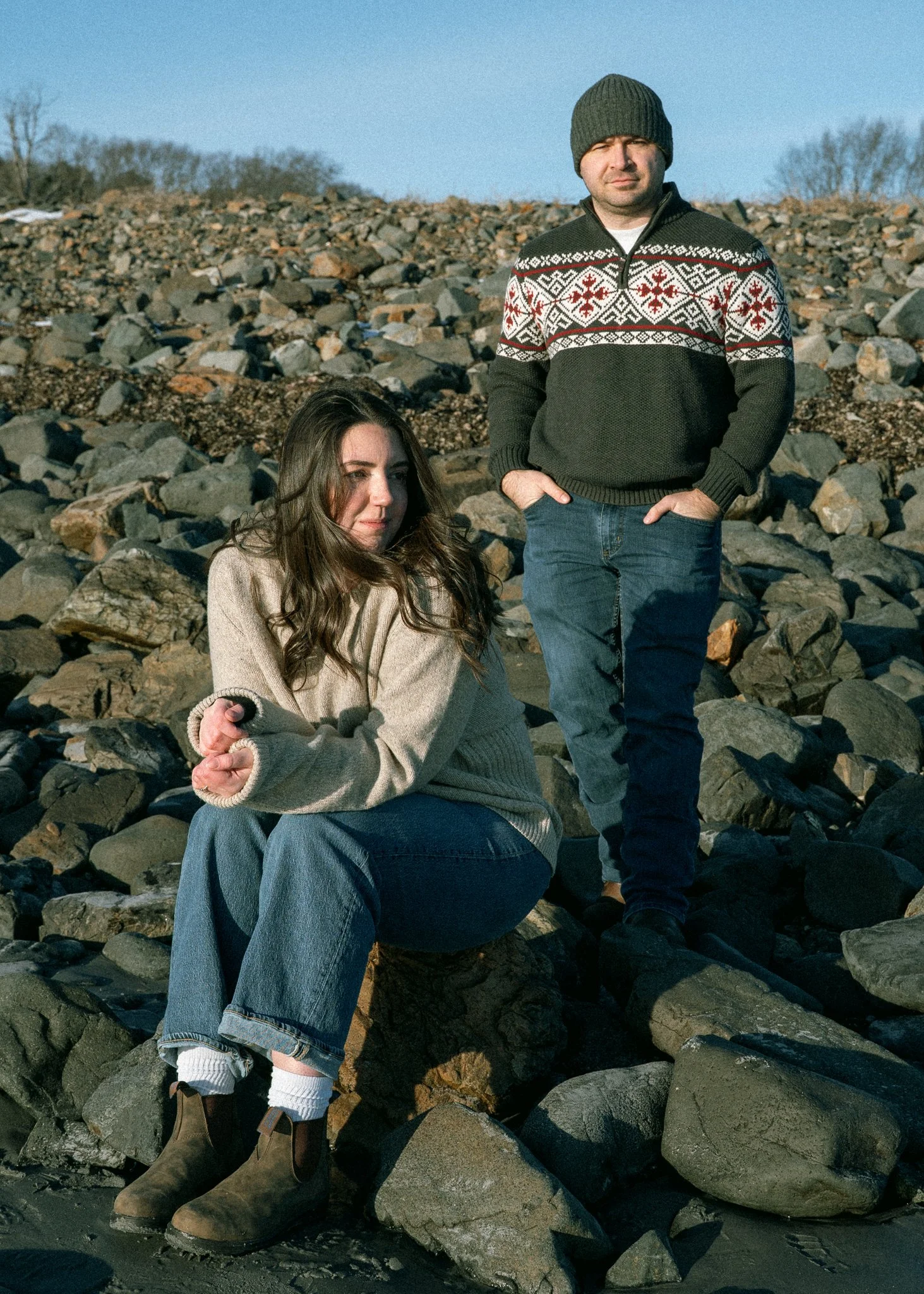 A man and a woman on a rocky beach, with the man standing and the woman sitting on a large rock, both dressed warmly, during daylight.
