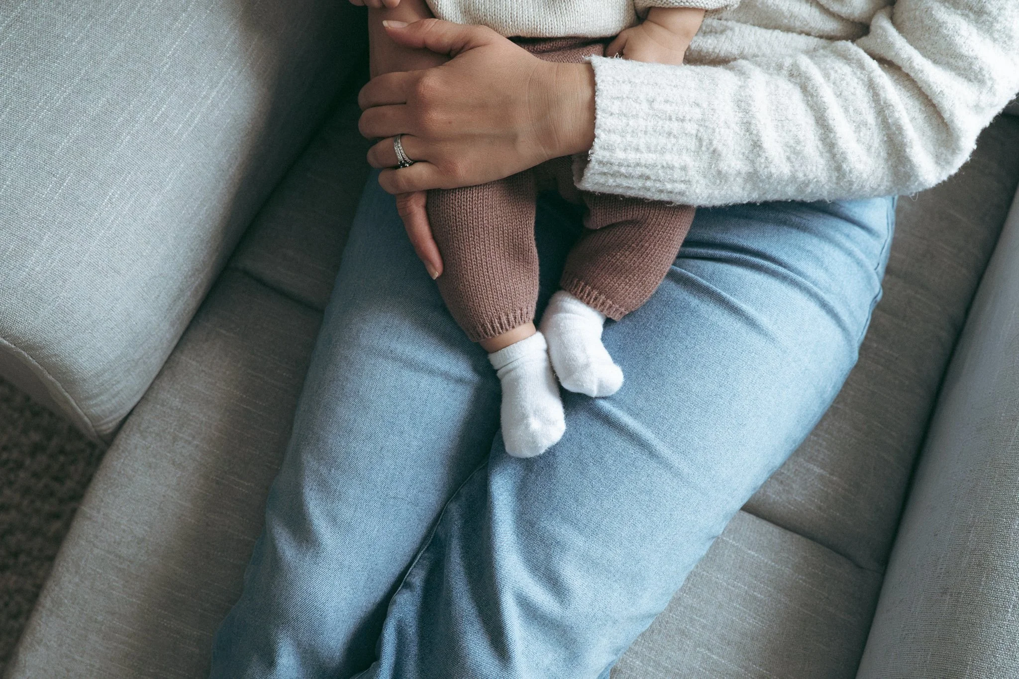 A woman sitting on a beige couch holding a small child who is wearing brown pants and white socks, with the woman's left hand resting on the child's leg.