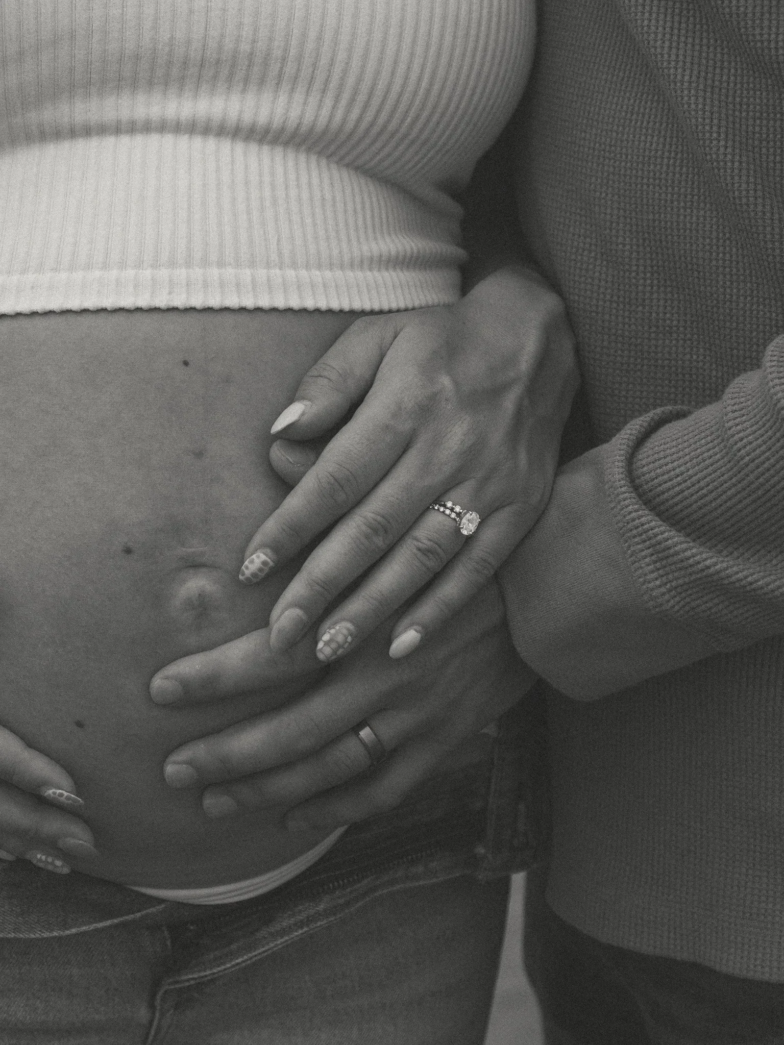 Close-up of a pregnant woman's belly with hands resting on it, wearing rings, with a man’s hand also on the belly. In black and white.