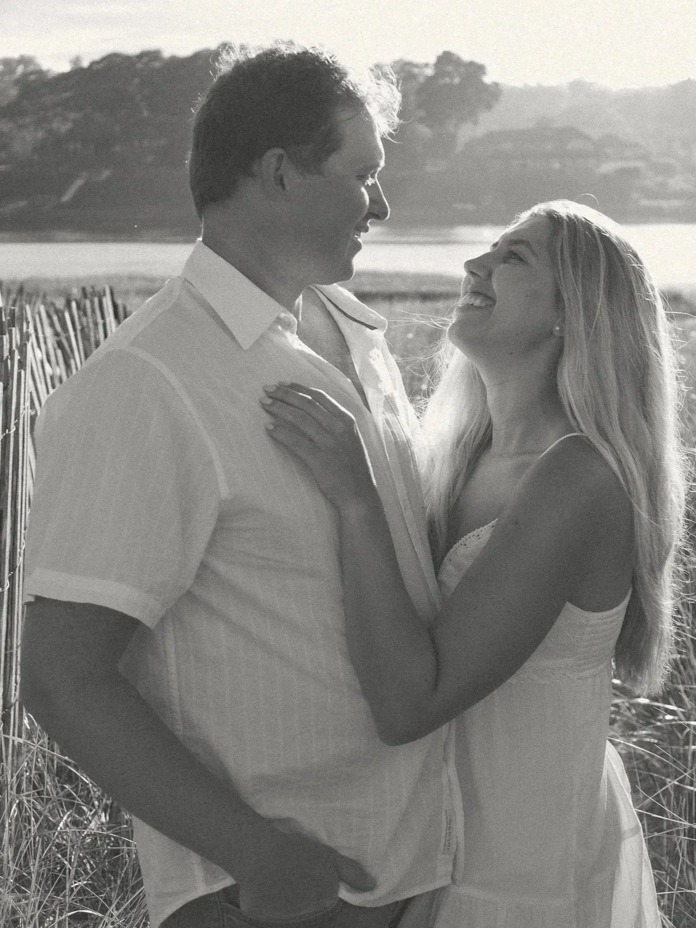 A black and white photo of a smiling man and woman looking at each other, standing near a body of water with a landscape of trees and hills in the background.